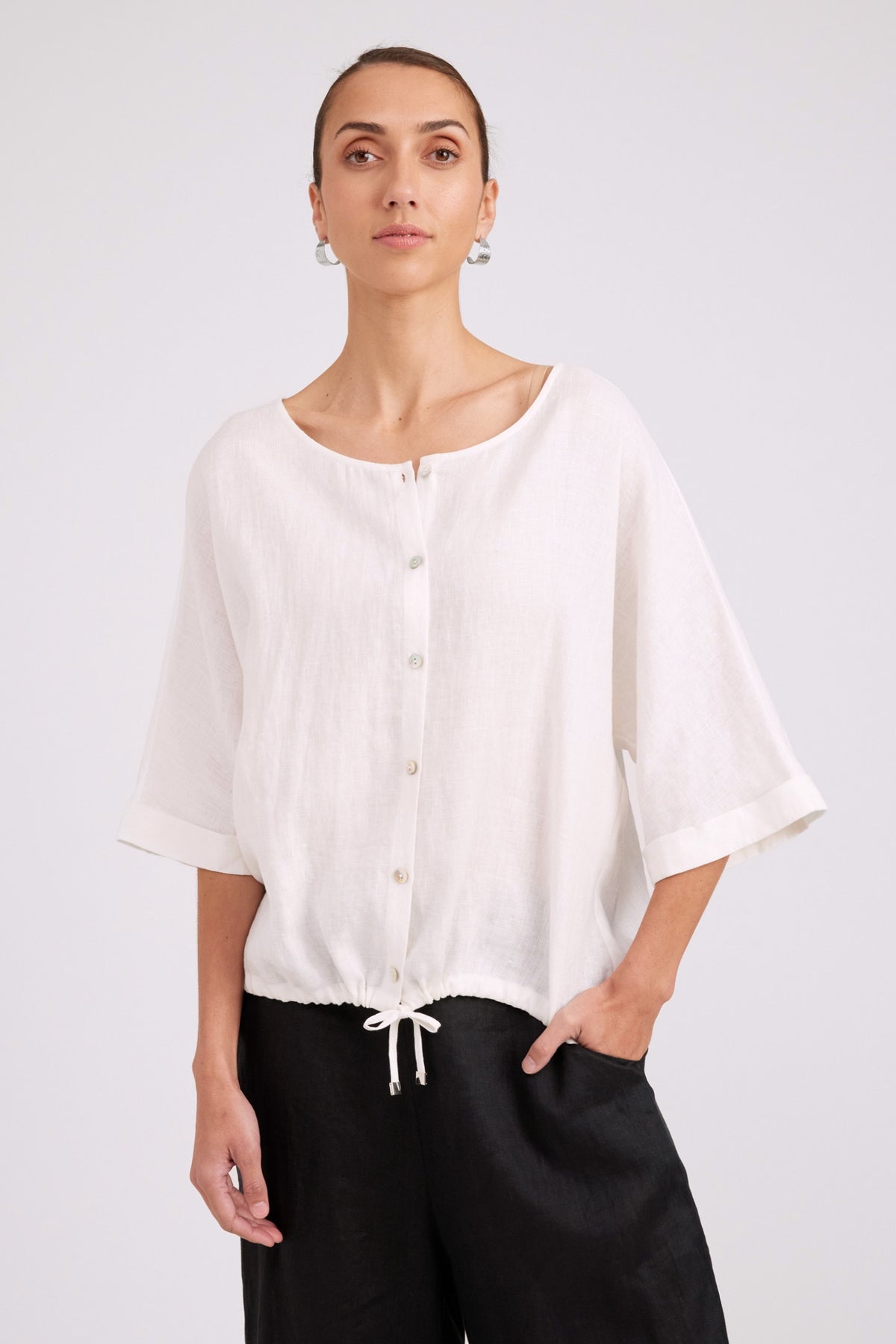 A woman wearing a loose white short-sleeve button-front blouse with a drawstring hem, paired with black pants, photographed against a light gray studio background.