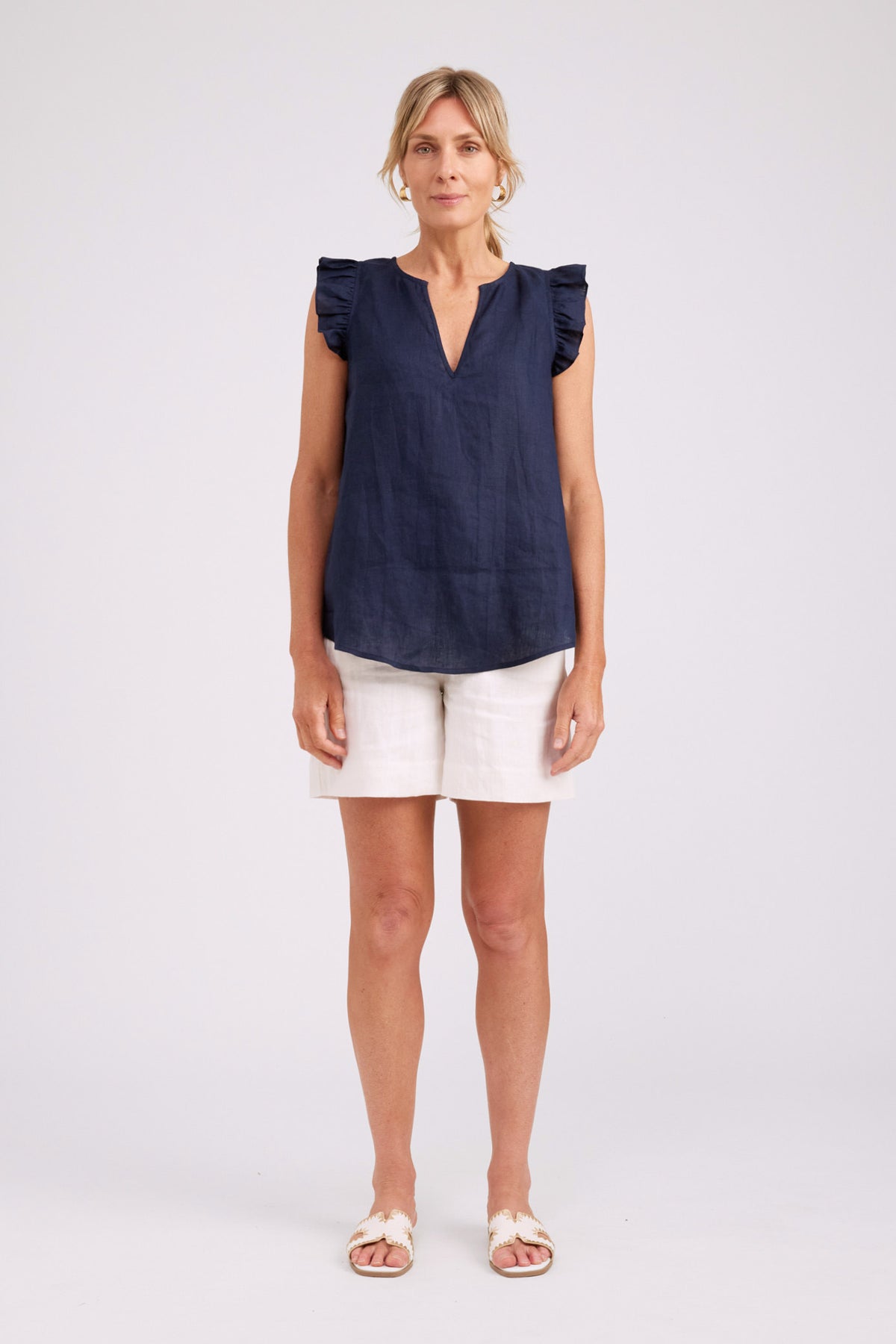 A woman modeling a sleeveless navy blue top with ruffled shoulders and a split V-neckline, paired with white shorts and white sandals, against a light gray studio background.