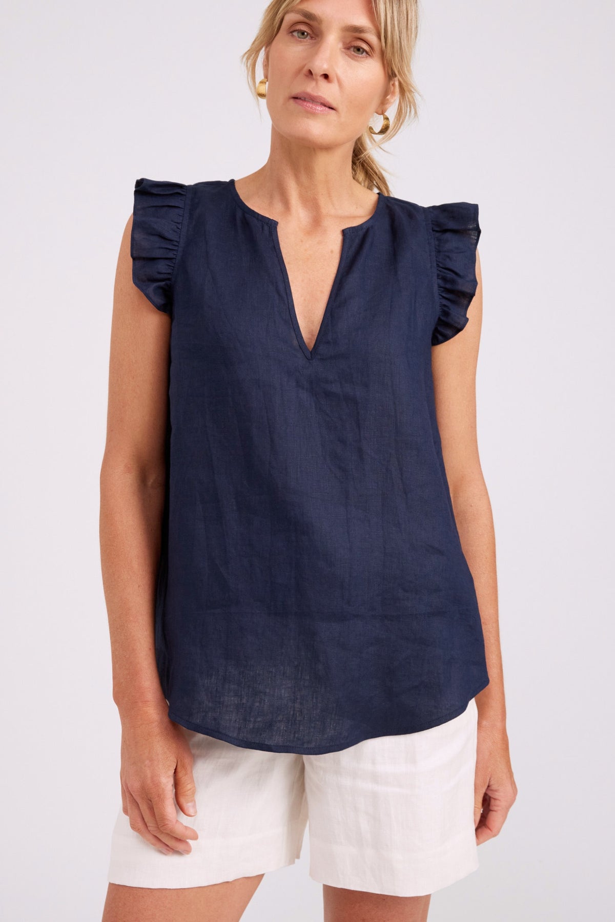 A woman wearing a navy blue sleeveless top with ruffled cap sleeves and a split V-neckline, paired with white shorts, against a plain light gray studio background.