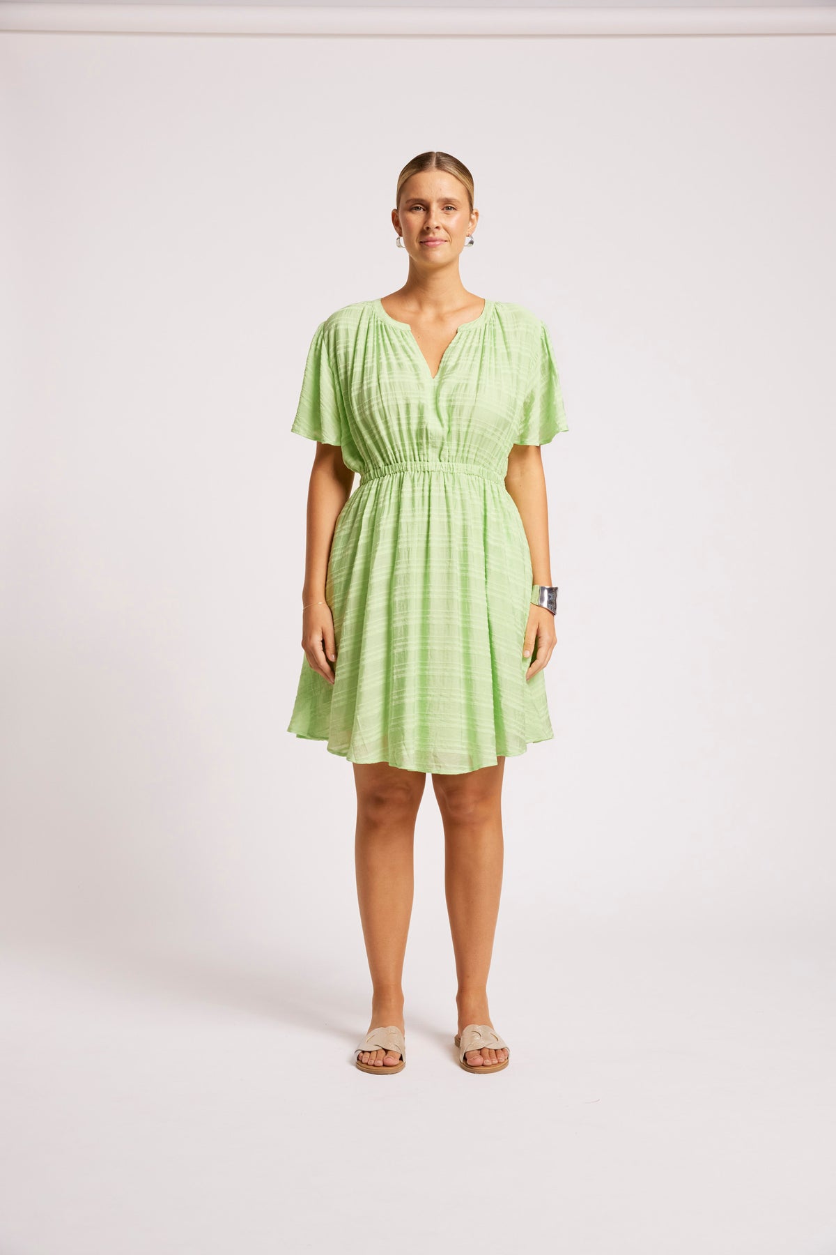 A woman modeling a light green textured mini dress with short flutter sleeves, a gathered waist, and a V-neckline, standing against a plain light gray studio background.