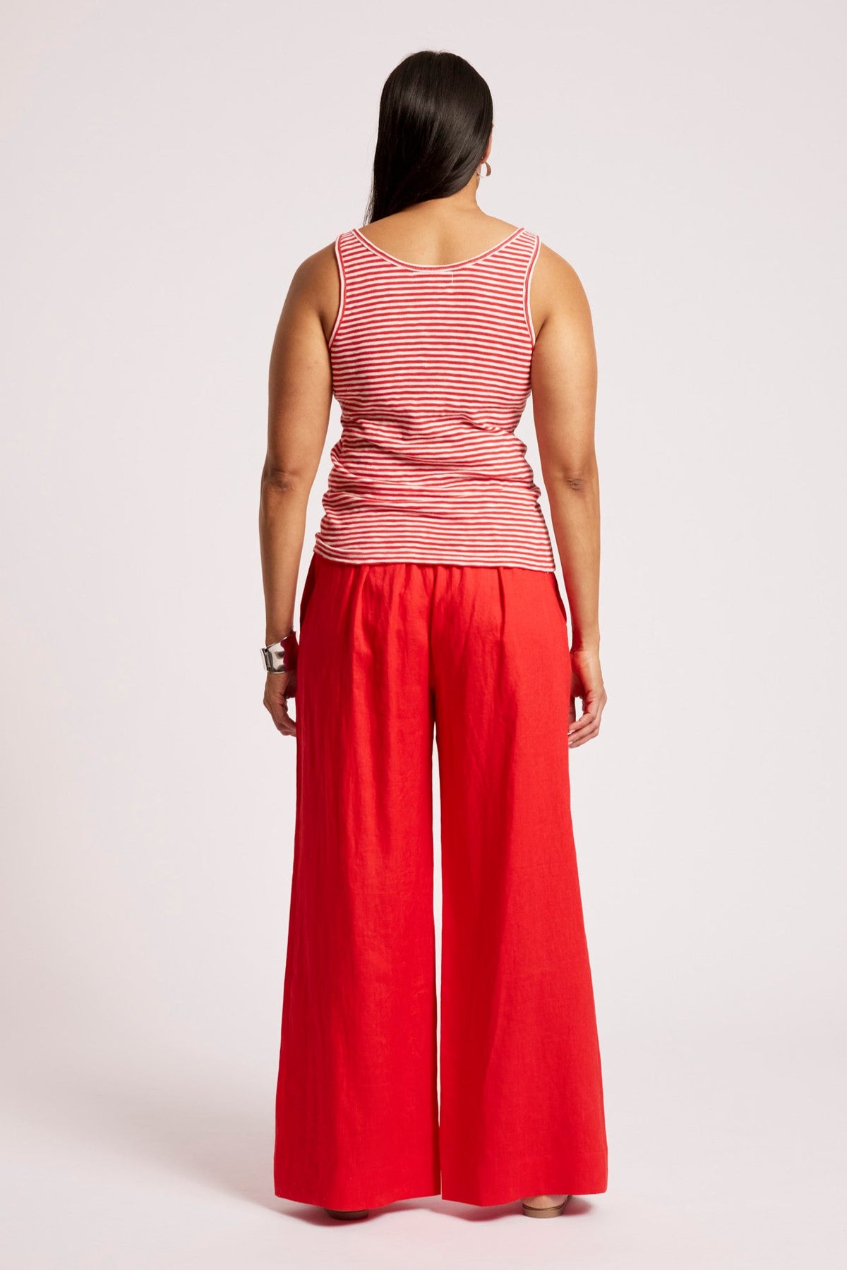 A woman viewed from the back wearing a sleeveless red and white striped top and bright red wide-leg pants, standing against a light gray studio background.