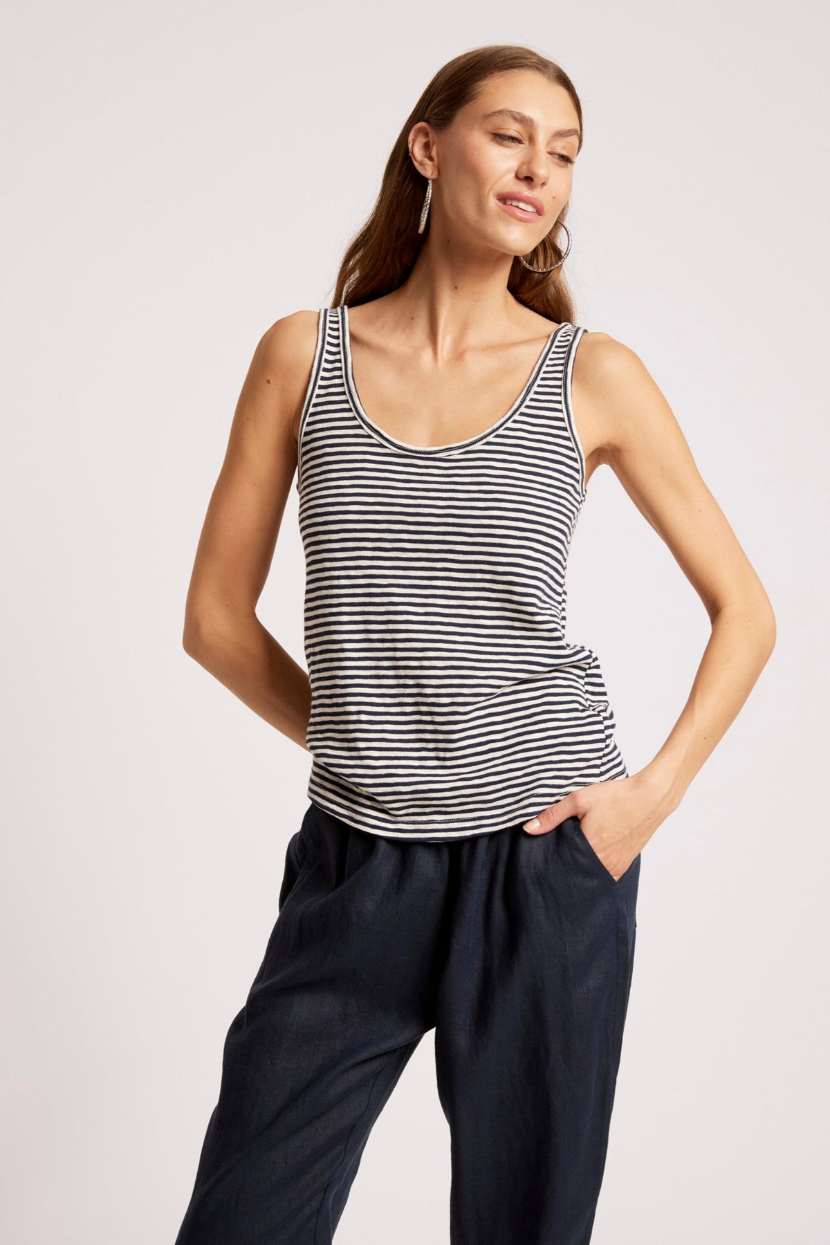 A woman modeling a sleeveless tank top with thin black and white horizontal stripes, paired with dark navy pants, against a plain light gray studio background.