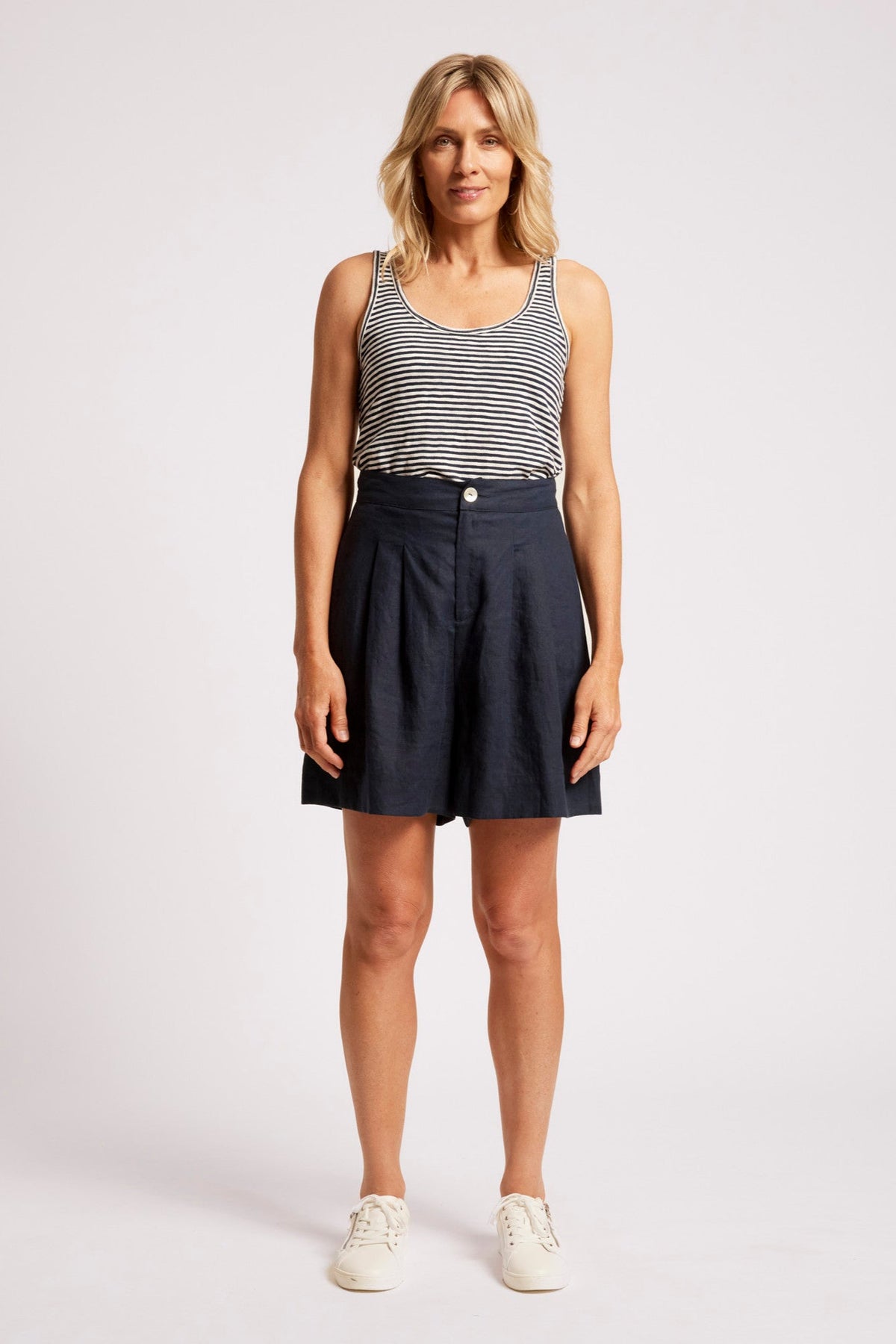 A woman modeling a sleeveless black and white striped tank top paired with dark navy blue pleated shorts and white sneakers, standing against a light gray studio background.