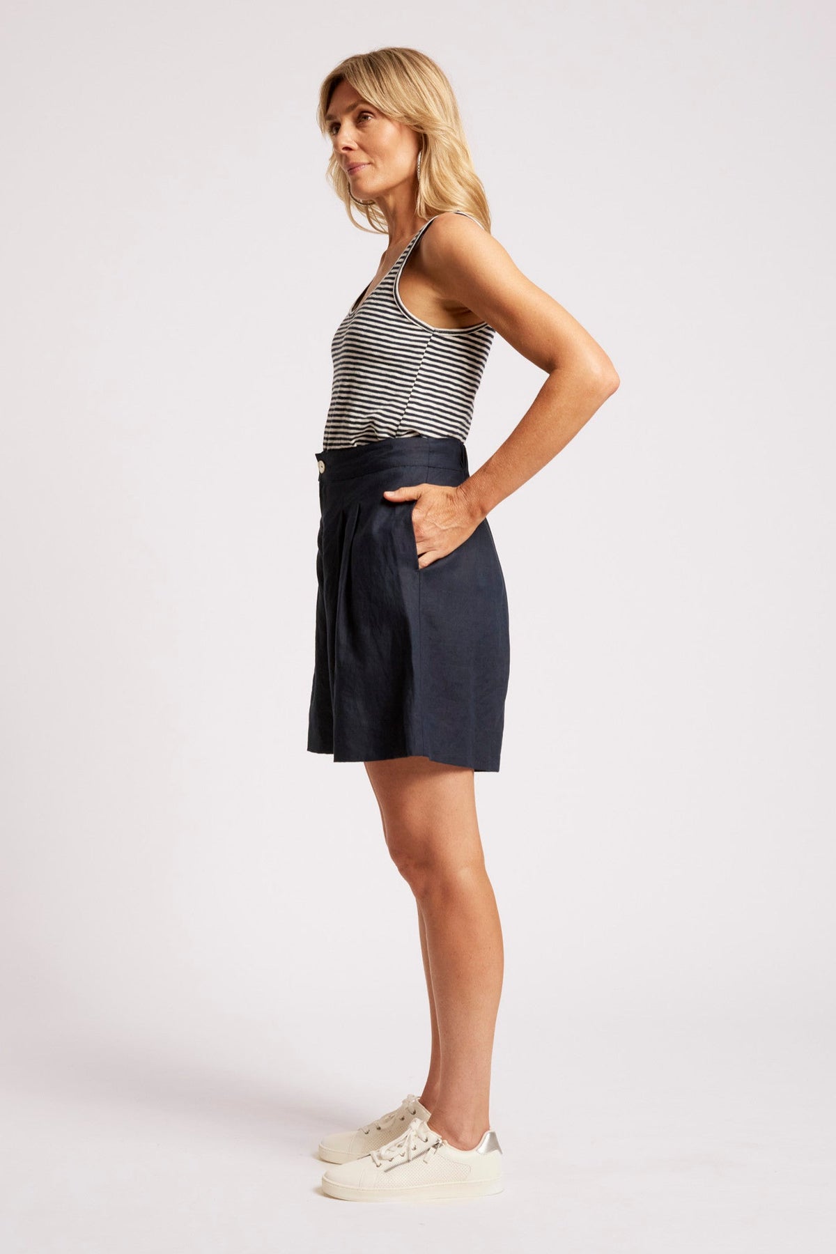A woman modeling a pair of dark navy blue high-waisted shorts with a striped tank top and white sneakers, standing against a light gray studio background.