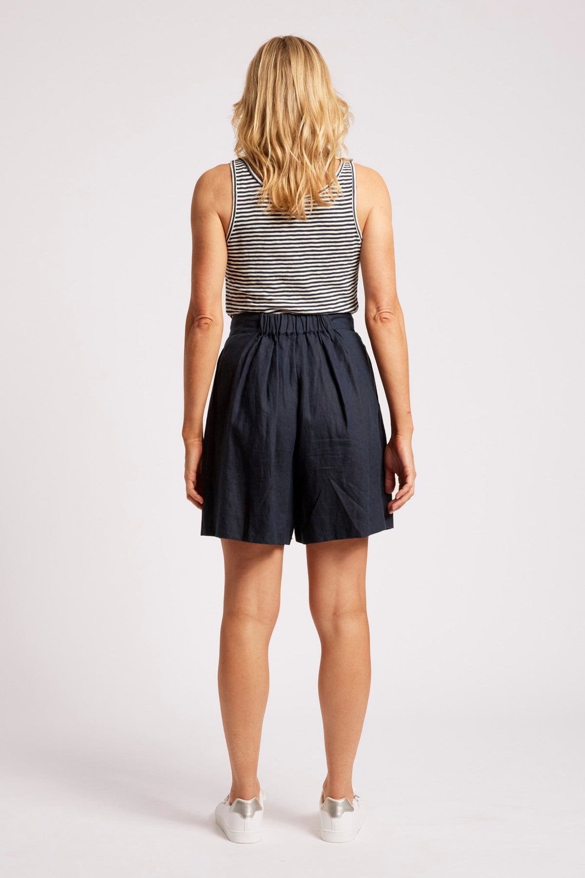 A woman viewed from behind wearing a sleeveless black and white striped top and dark navy blue high-waisted shorts, paired with white sneakers, against a light gray studio background.
