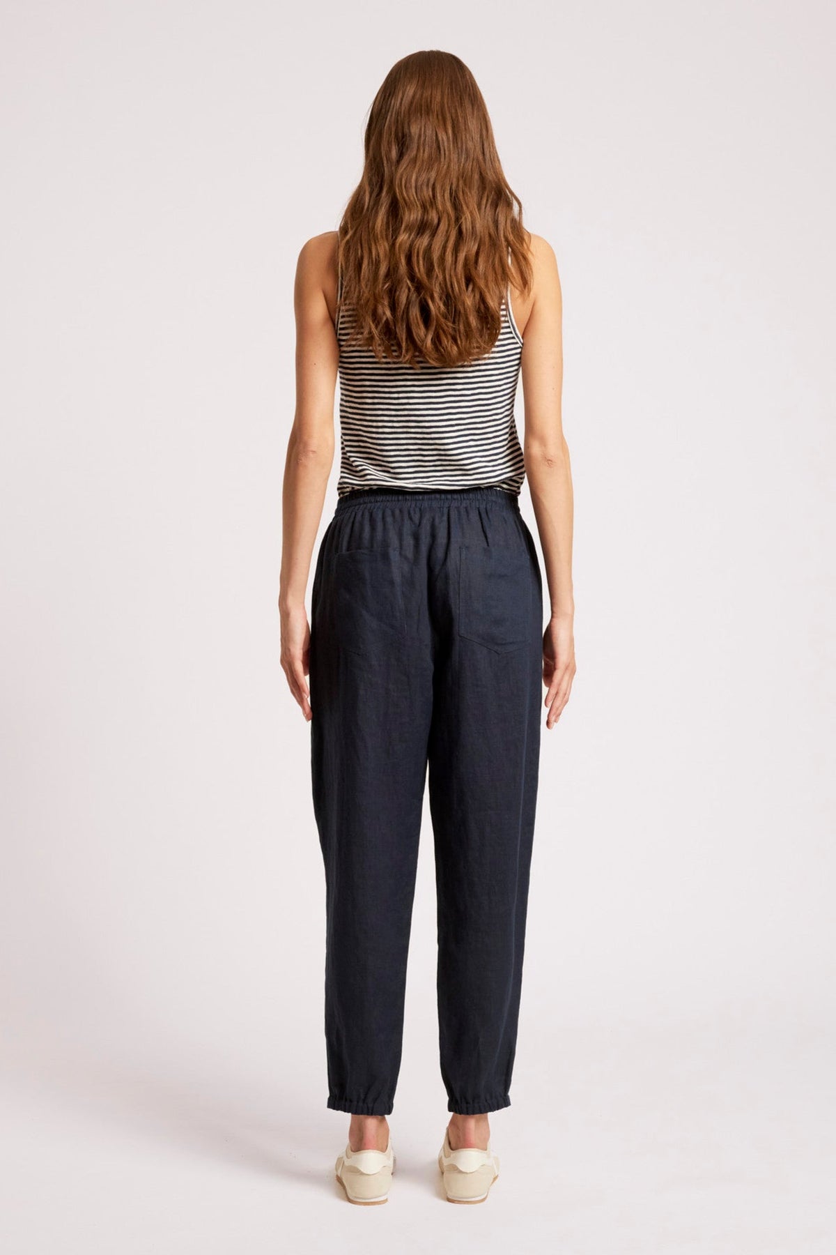 A woman viewed from behind wearing dark navy blue linen trousers, a black and white striped tank top, and cream-colored shoes, set against a plain light gray studio background.