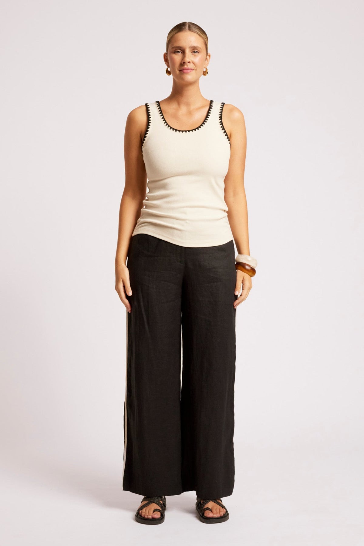A woman modeling a cream ribbed tank top with black trim and black wide-leg pants against a light gray studio background.