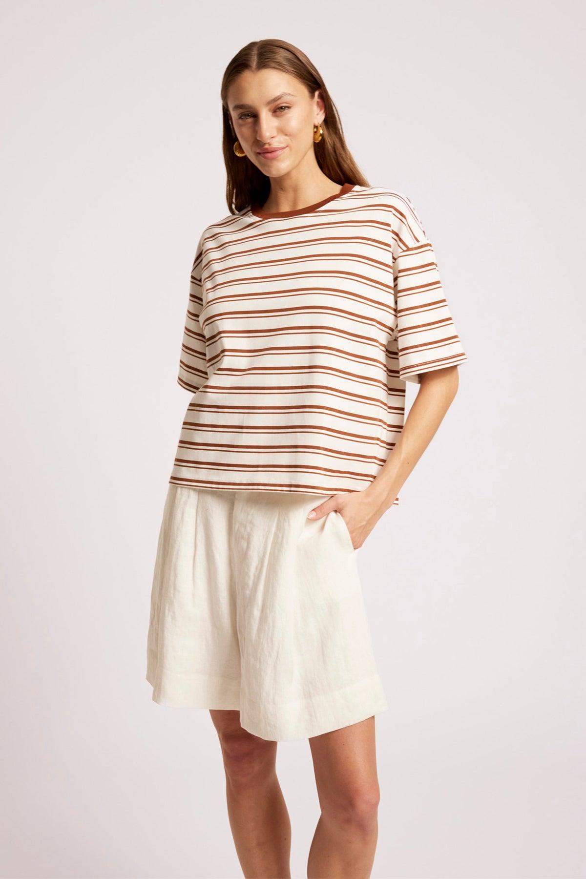 A woman modeling a short-sleeve crewneck top with thin brown and beige horizontal stripes, paired with an off-white linen skirt, against a light gray studio background.