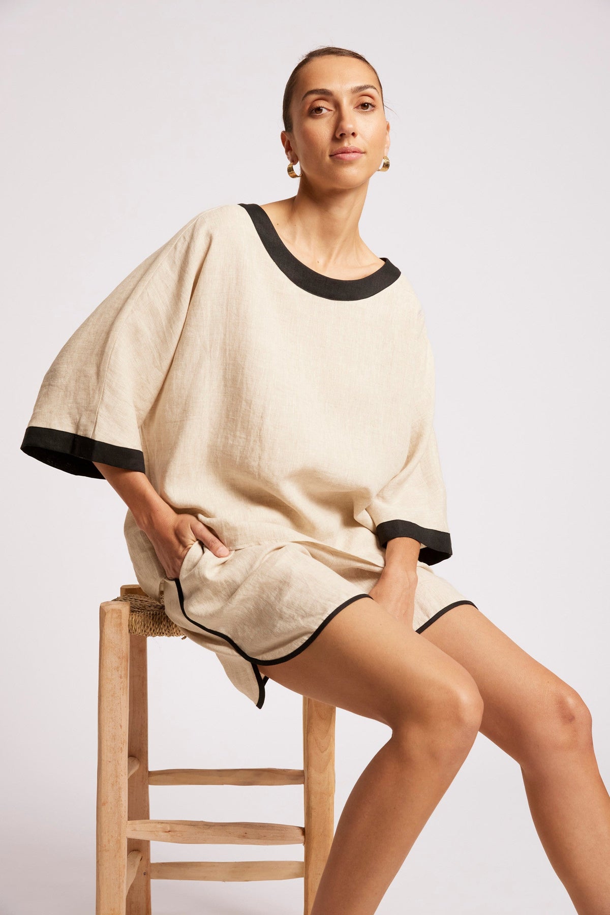 A woman seated on a wooden stool wearing a loose beige linen-like short dress or tunic with black trim at the neckline, sleeve cuffs, and hem, against a light gray studio background.