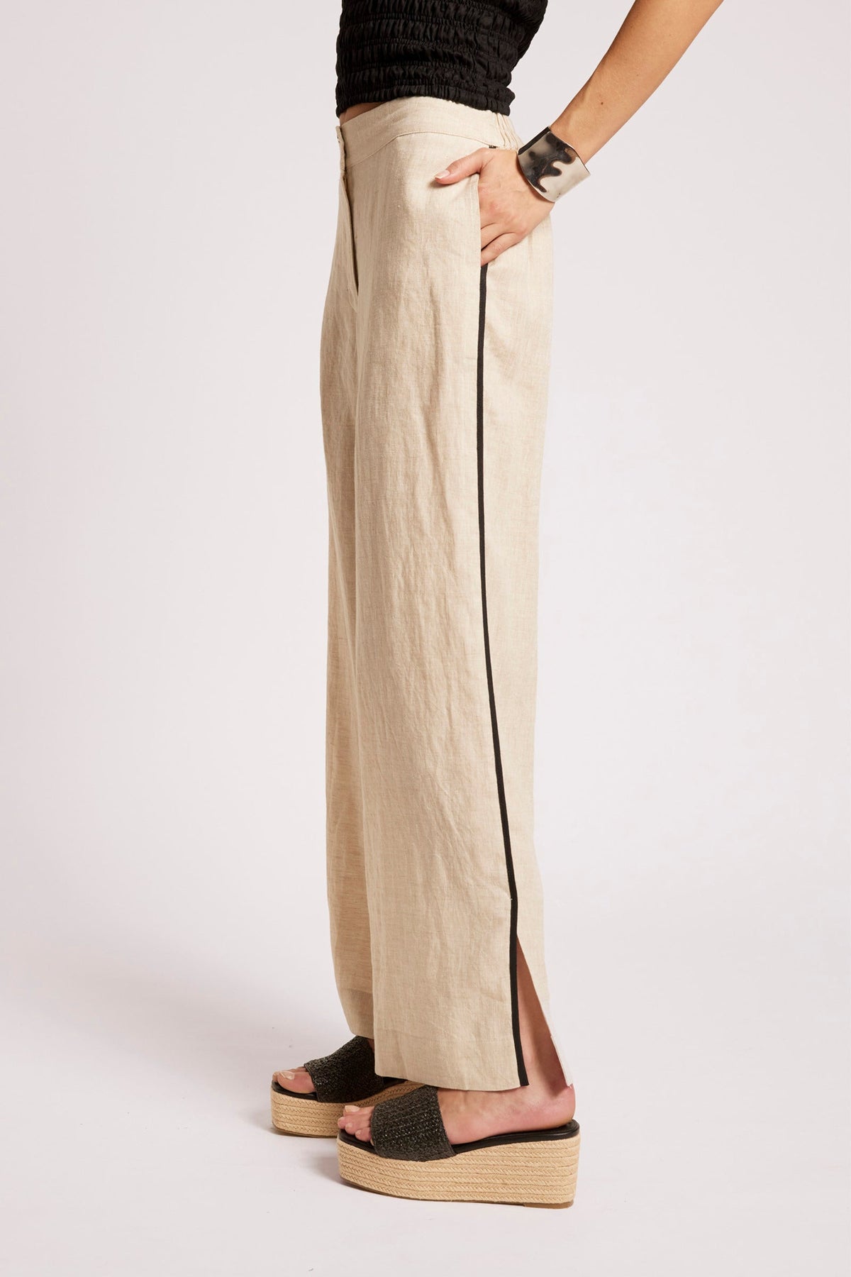 A person wearing wide-leg beige trousers with a thin black side stripe, paired with black platform slide sandals, shown from the waist down against a plain light background.