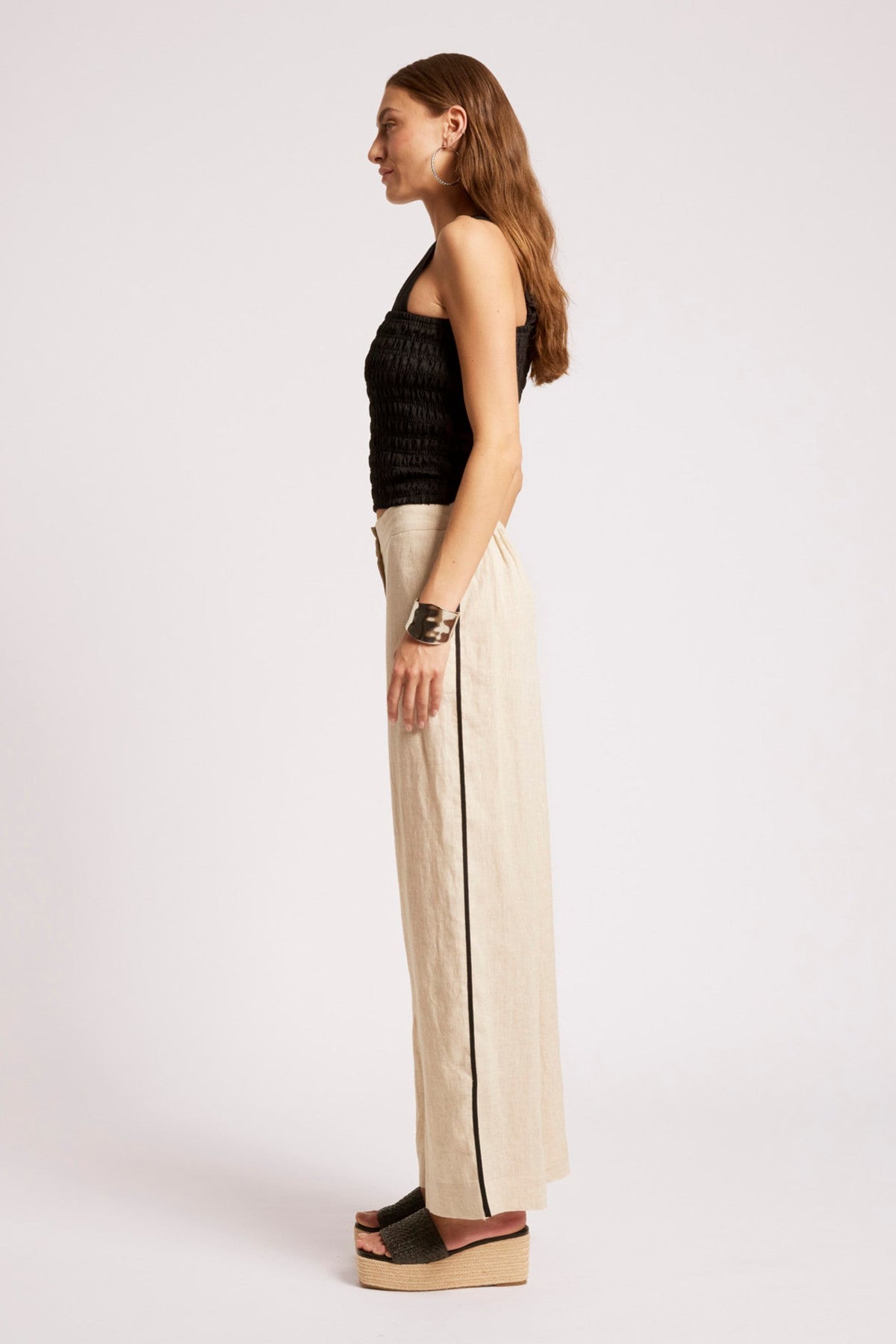 A woman stands in profile wearing a black ribbed halter top, wide-leg cream trousers with a thin black side stripe, and brown platform sandals against a plain light gray studio background.