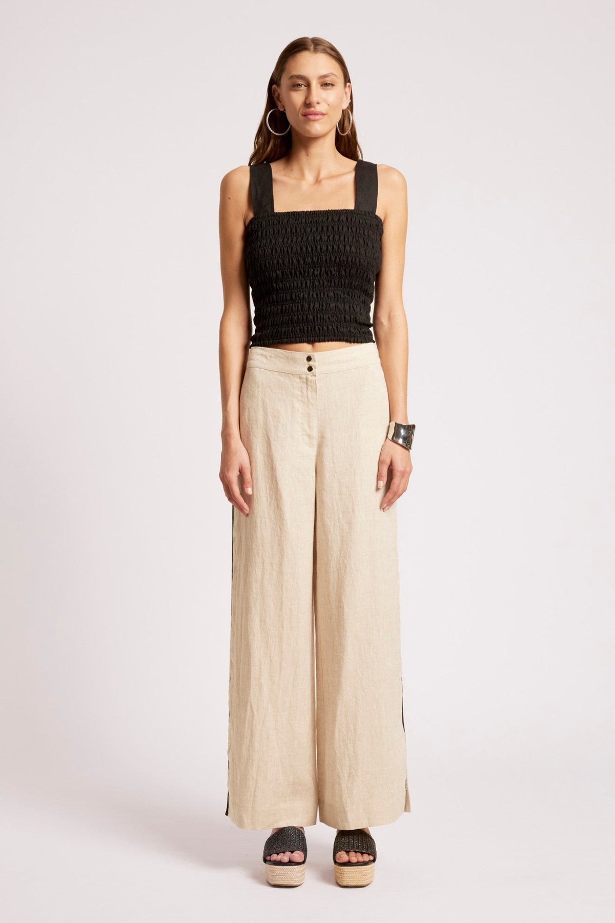 A woman modeling a black smocked sleeveless crop top and beige wide-leg trousers with side buttons, paired with black platform sandals, against a light gray studio background.