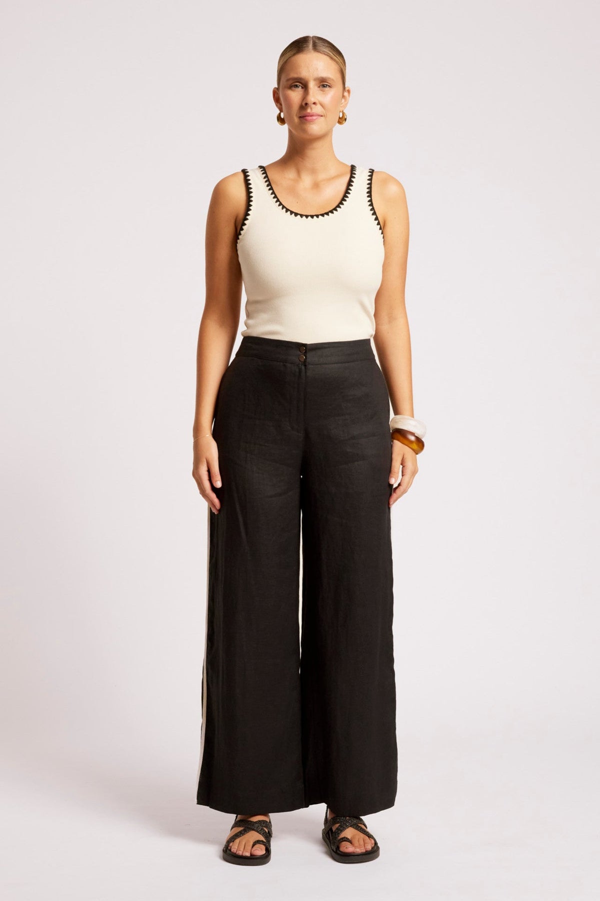 A woman stands against a light gray studio background wearing a cream sleeveless top with black trim and black wide-leg pants.