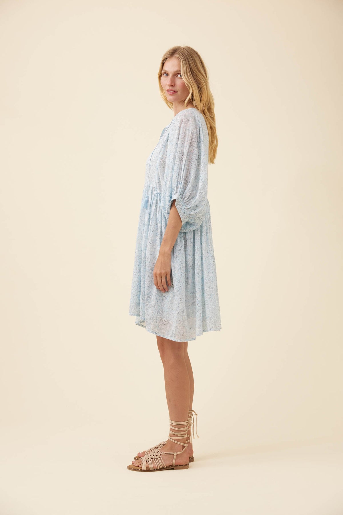 A woman modeling a light blue patterned midi dress with three-quarter sleeves, standing in profile against a solid cream background.