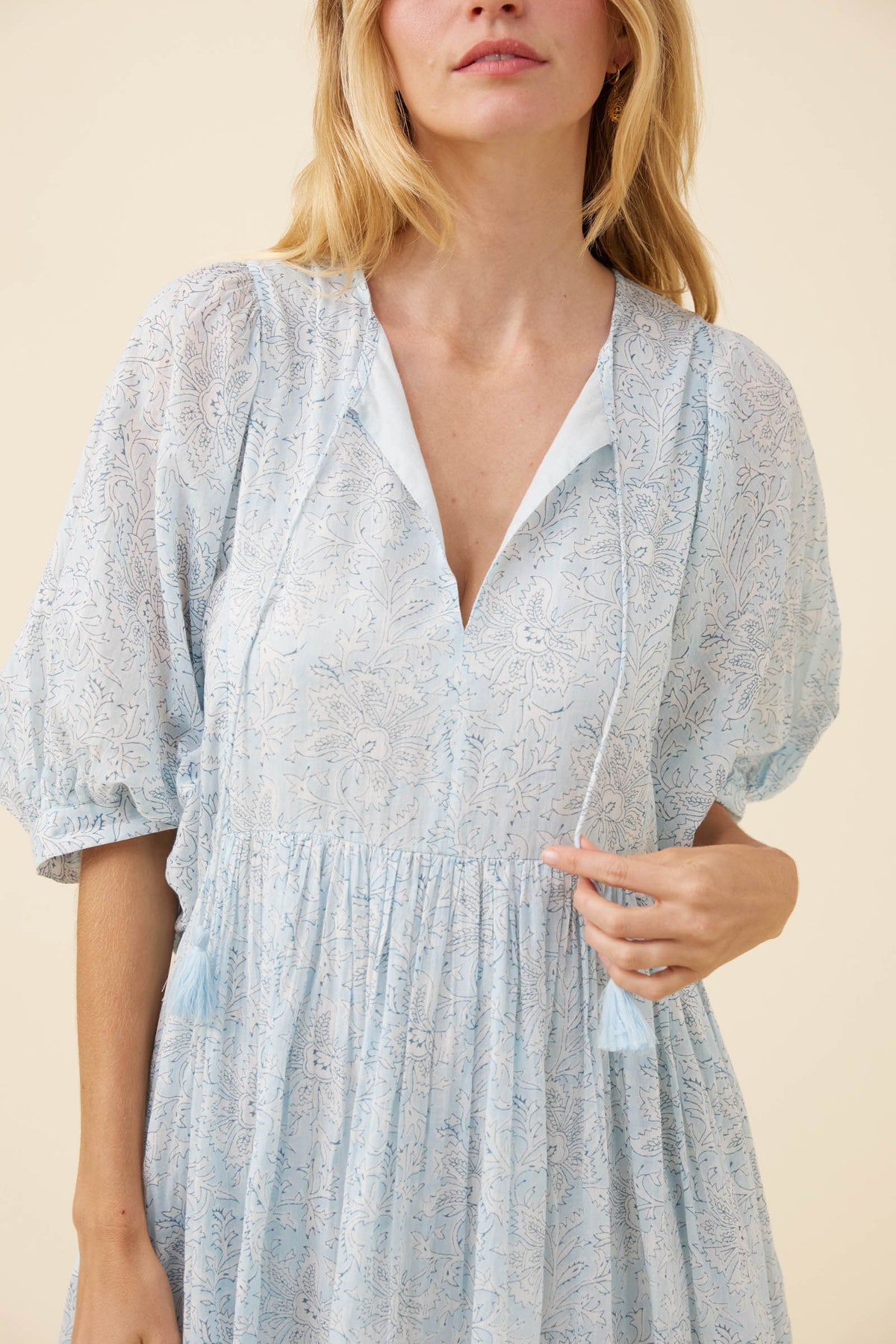 A woman wearing a light blue lace dress with puff sleeves, a deep V-neckline, and tassel ties, photographed against a solid beige background.