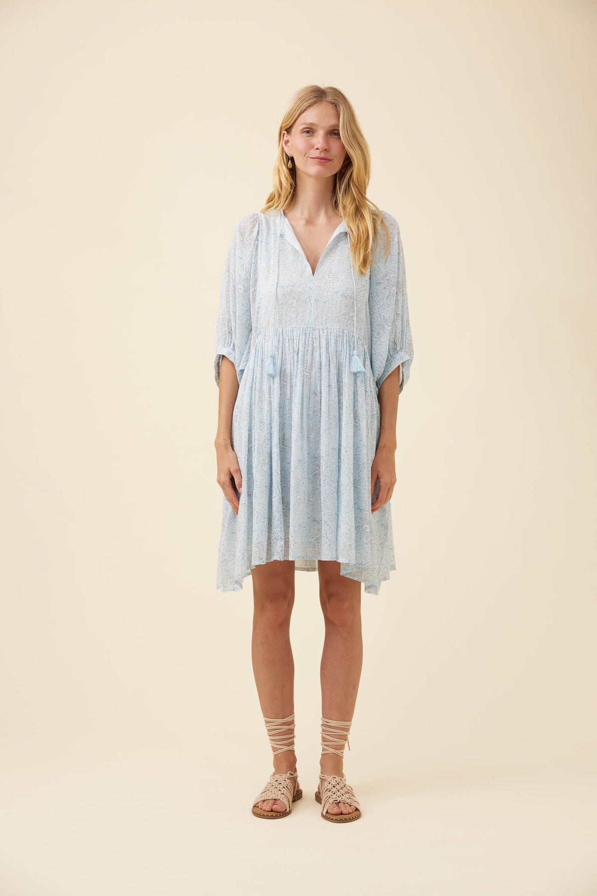 A woman modeling a light blue and white patterned short-sleeve dress with a V-neck and gathered waist, standing against a solid beige studio background.