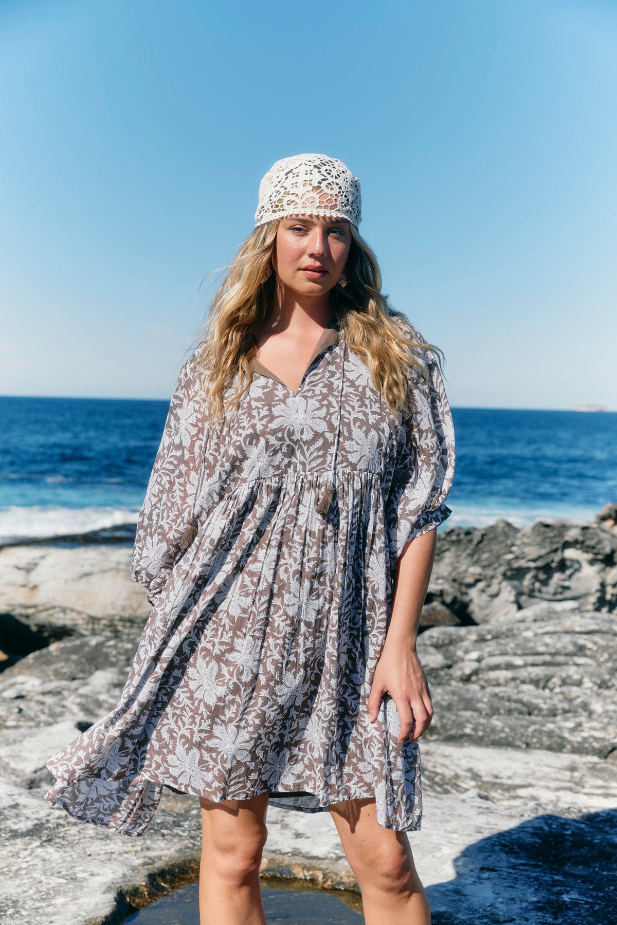 A woman wearing a loose, short-sleeve boho-style dress with a brown and white floral pattern, a white crochet headband, and standing on rocky shore with the ocean and blue sky in the background.