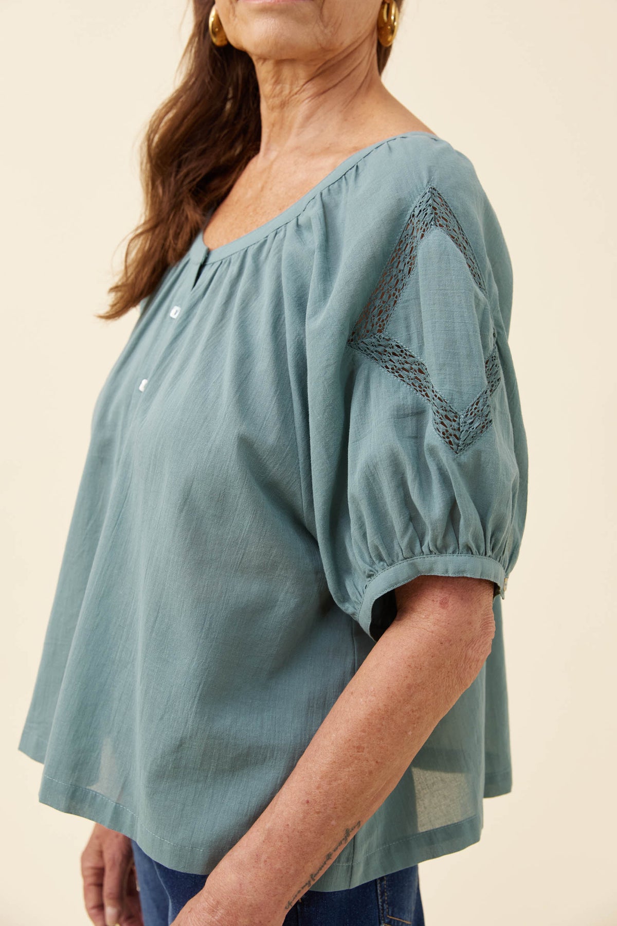 A woman wearing a dusty teal blue blouse with puff sleeves, a gathered neckline, and a geometric embroidered pattern on the upper sleeve, shown against a plain beige background.