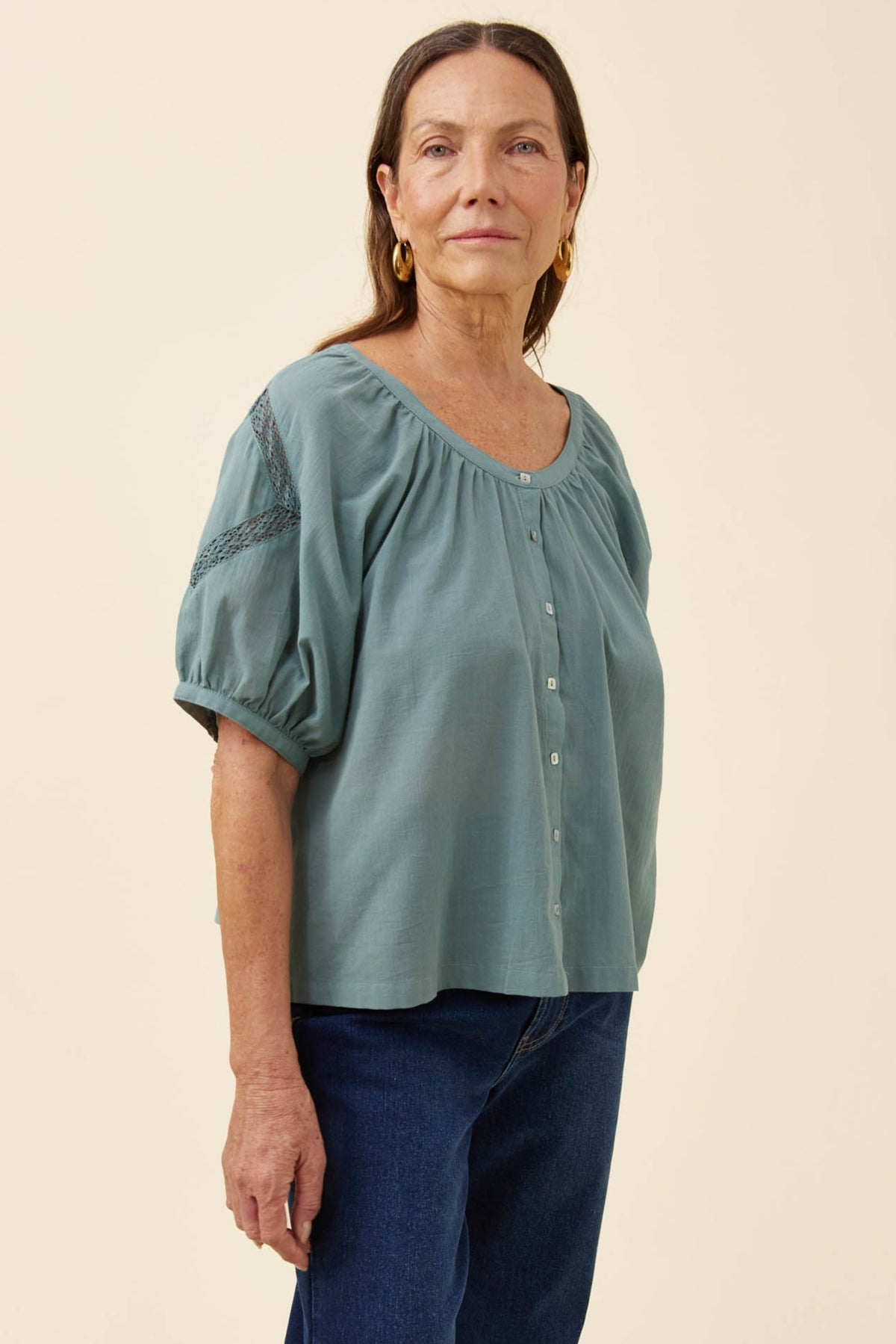 A woman modeling a sage green short-sleeve blouse with puff sleeves, a gathered neckline, and button-front closure, paired with dark blue jeans against a plain beige studio background.