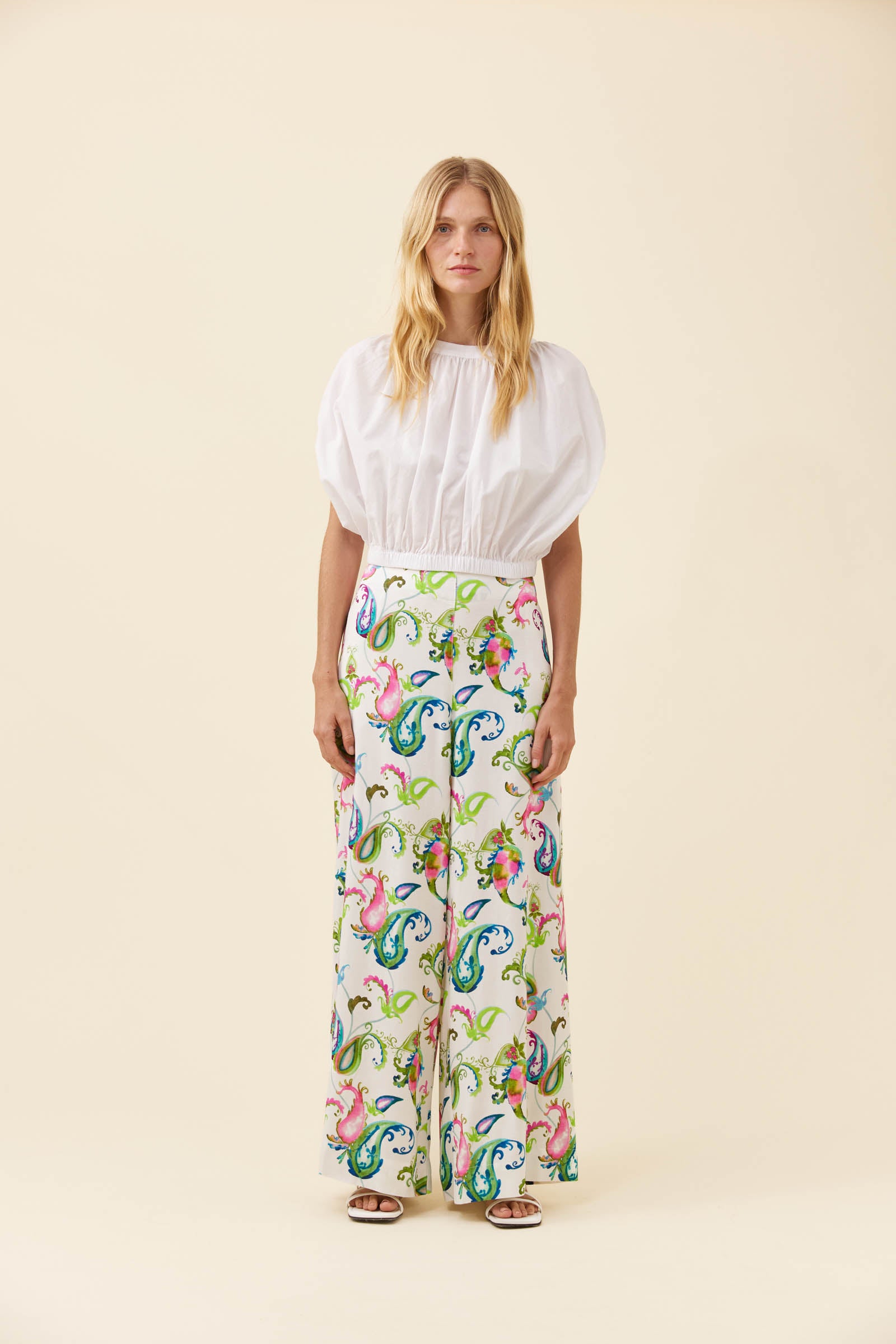 A woman stands against a plain beige studio background wearing a white puff-sleeve blouse and wide-leg trousers with a colorful paisley print.