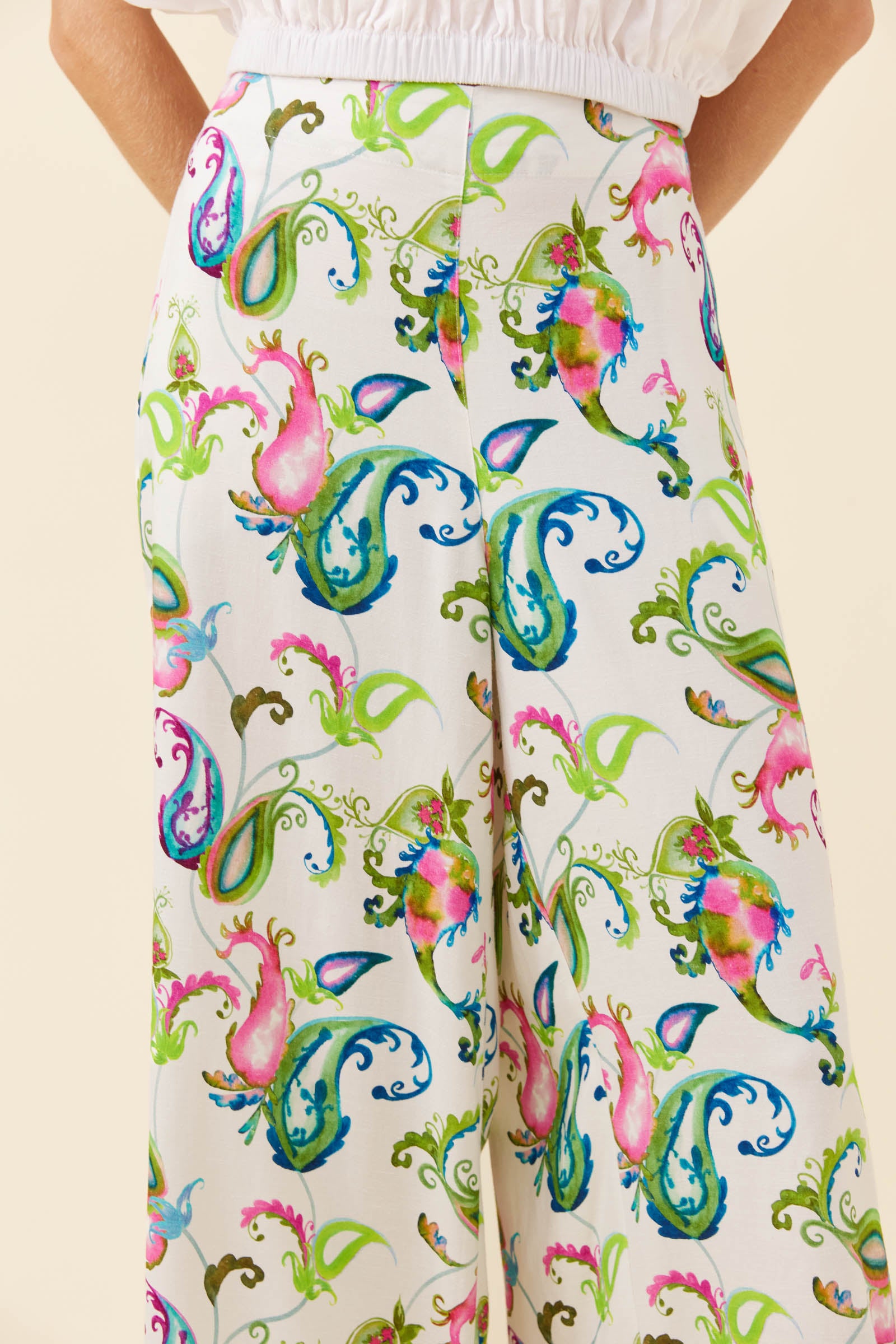 A close-up of a model wearing high-waisted, wide-leg trousers with a colorful paisley and floral print on an off-white base, paired with a white top.