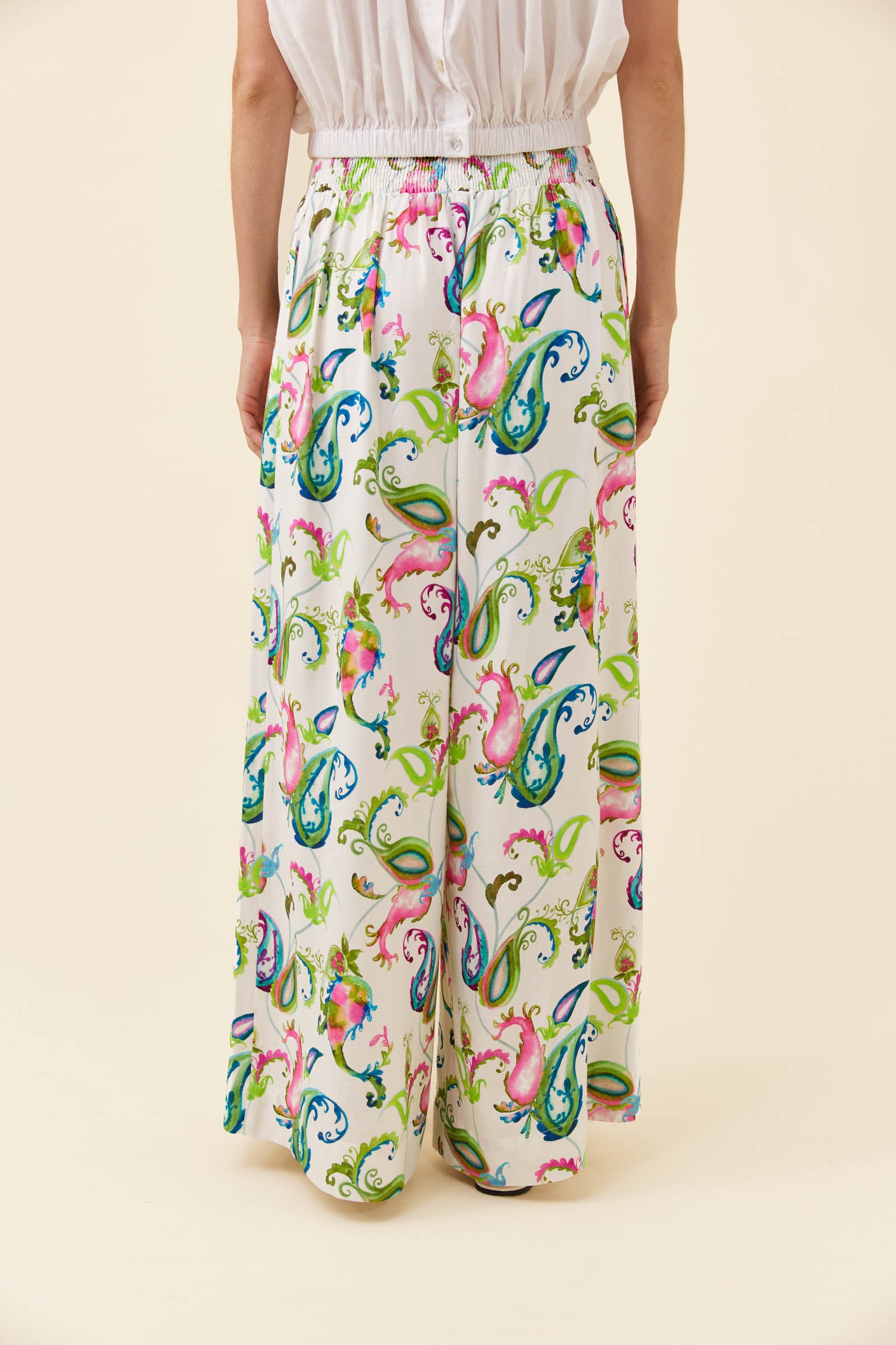 A model is shown from the waist down wearing high-waisted, wide-leg trousers with a vibrant multicolor paisley pattern on a cream background.