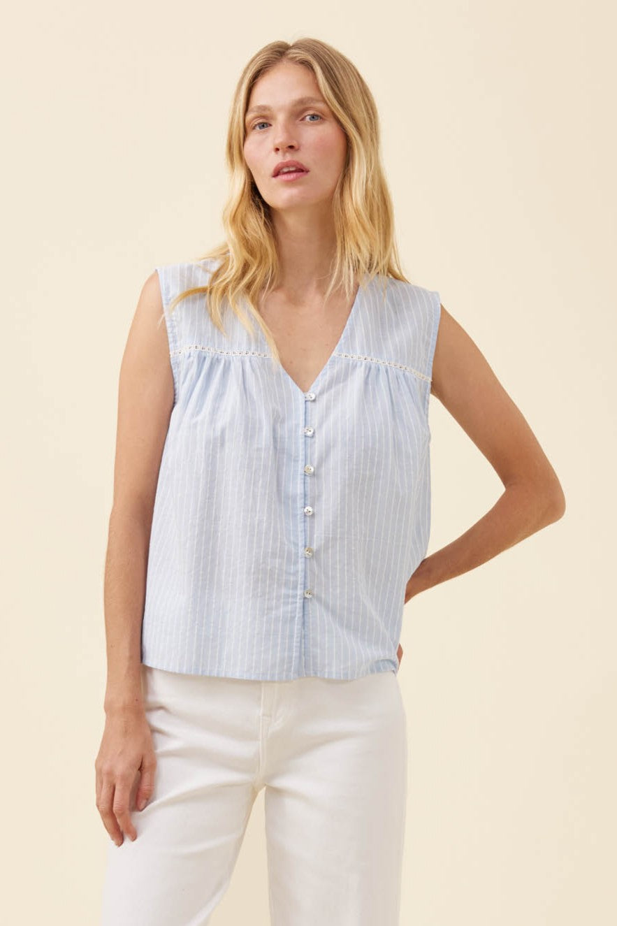 A woman modeling a sleeveless light blue and white vertically striped blouse with a V-neck, button front, and gathered shoulders, paired with white pants against a plain beige studio background.