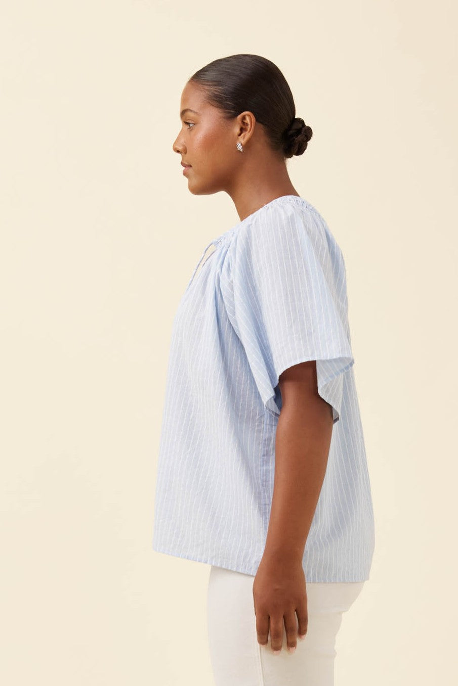 A woman shown in side profile wearing a light blue and white vertically striped short-sleeve blouse with a gathered neckline, paired with white pants, against a solid cream background.