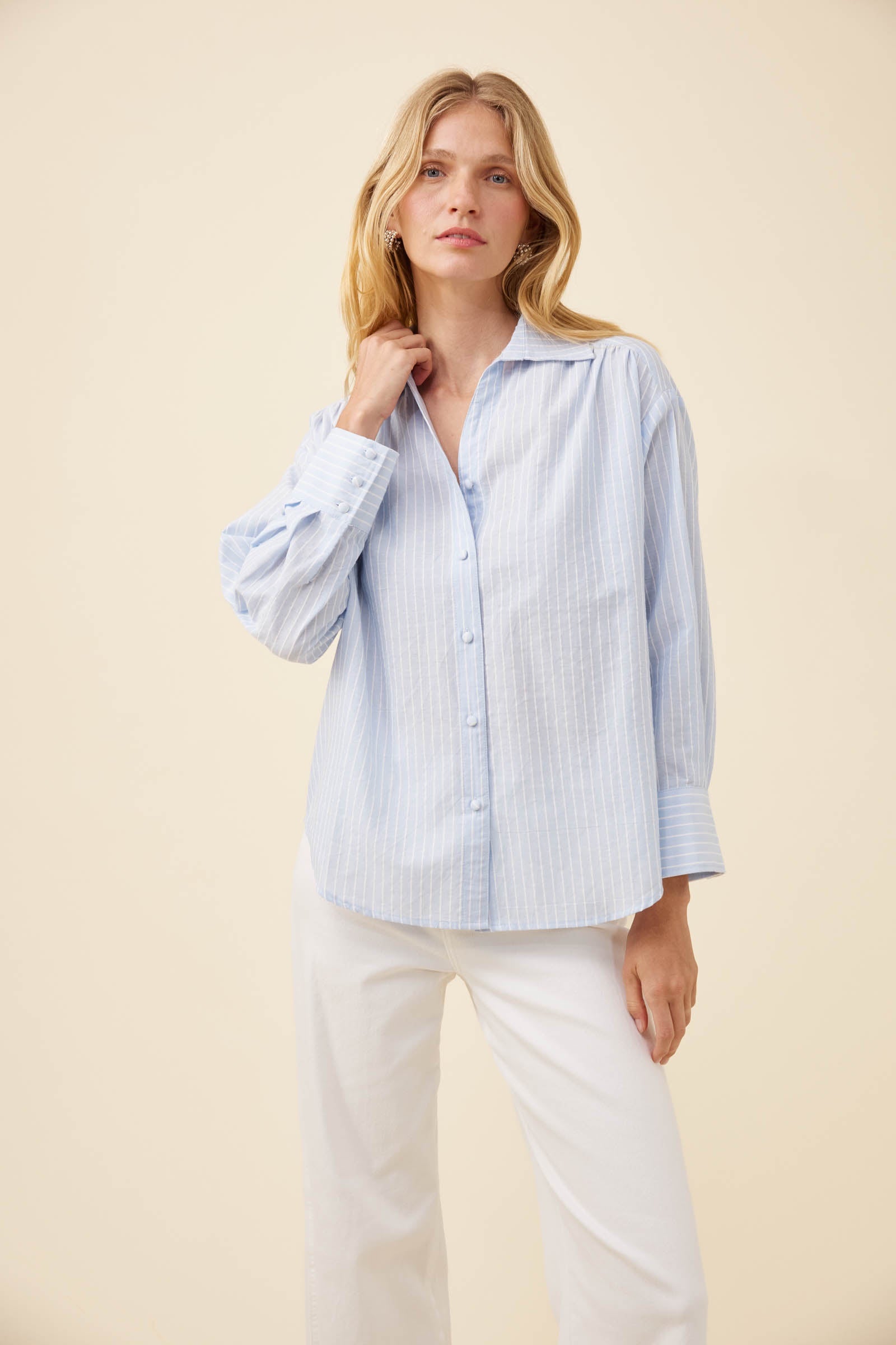 A woman modeling a light blue and white vertically striped button-down shirt with long sleeves, paired with white pants, against a solid beige studio background.