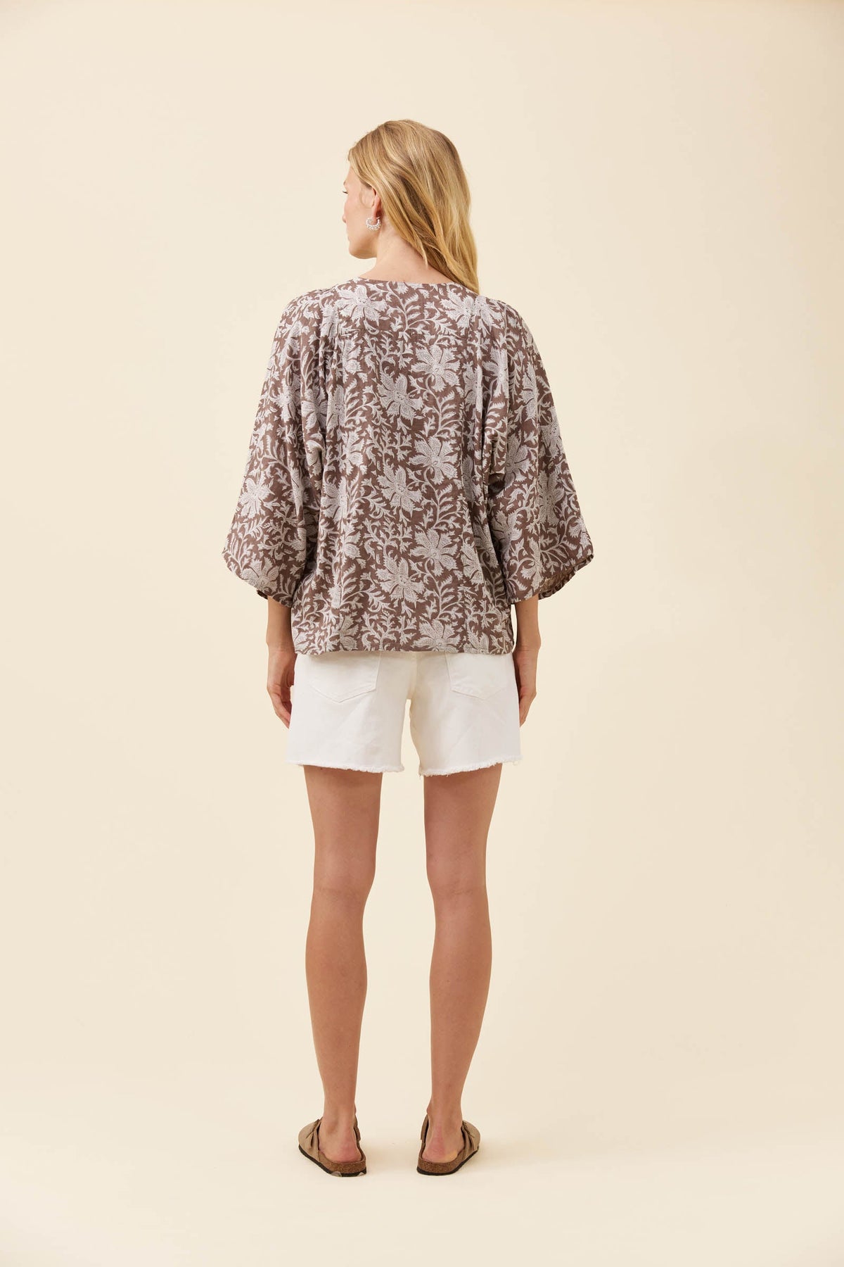A woman viewed from the back wearing a loose, short-sleeve patterned top in brown and beige, paired with white shorts and brown sandals, against a solid cream background.