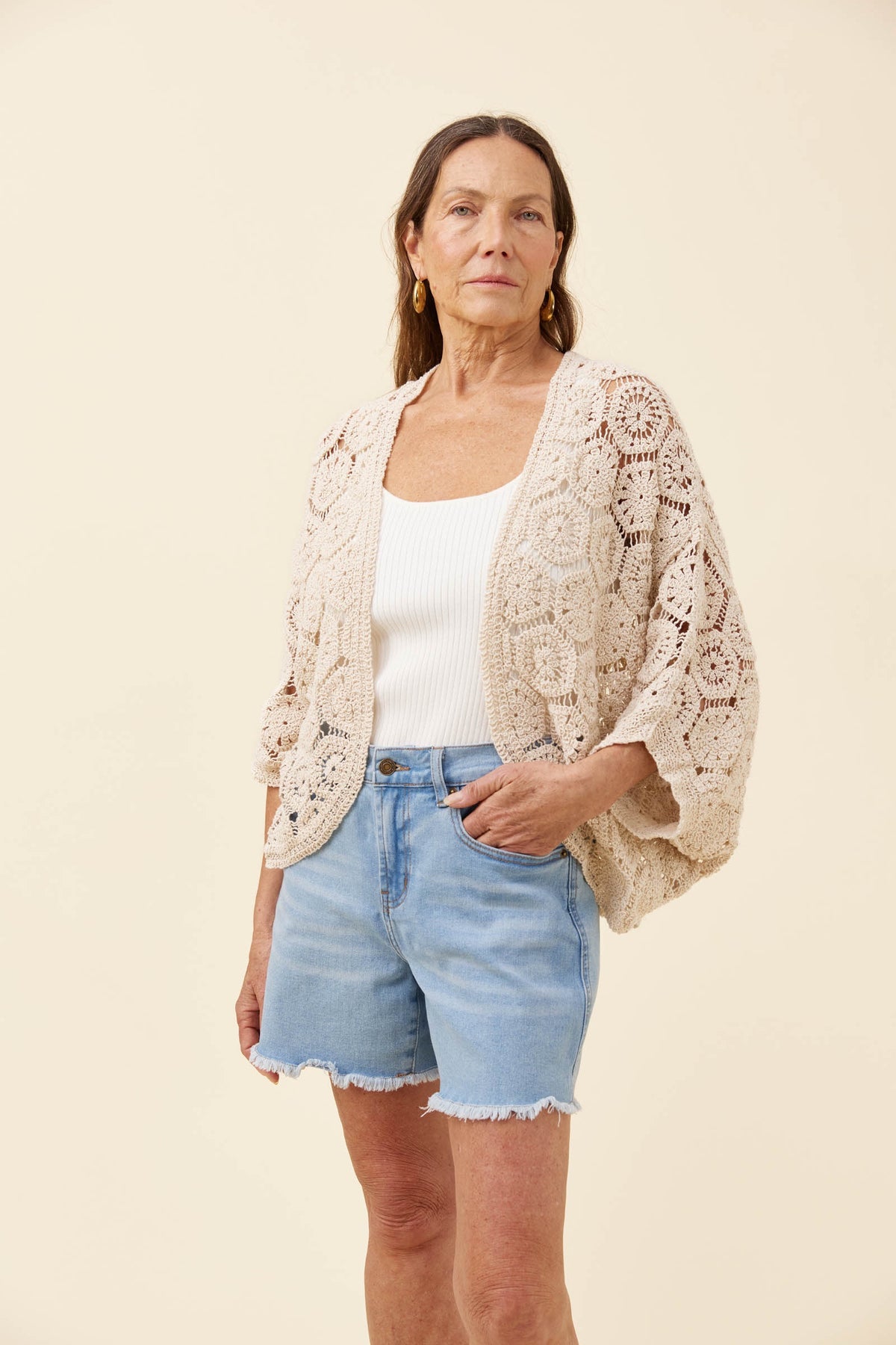A woman modeling a cream-colored crochet cardigan over a white tank top and light blue denim shorts, standing against a solid light beige background.