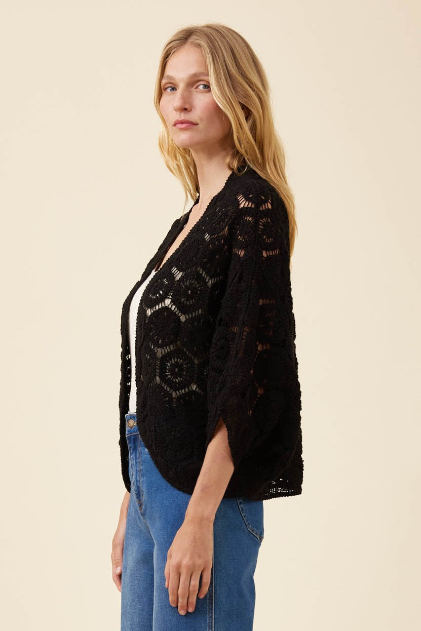 A woman wearing a black open-front crochet cardigan with three-quarter sleeves over a white top and blue jeans, photographed against a plain beige studio background.