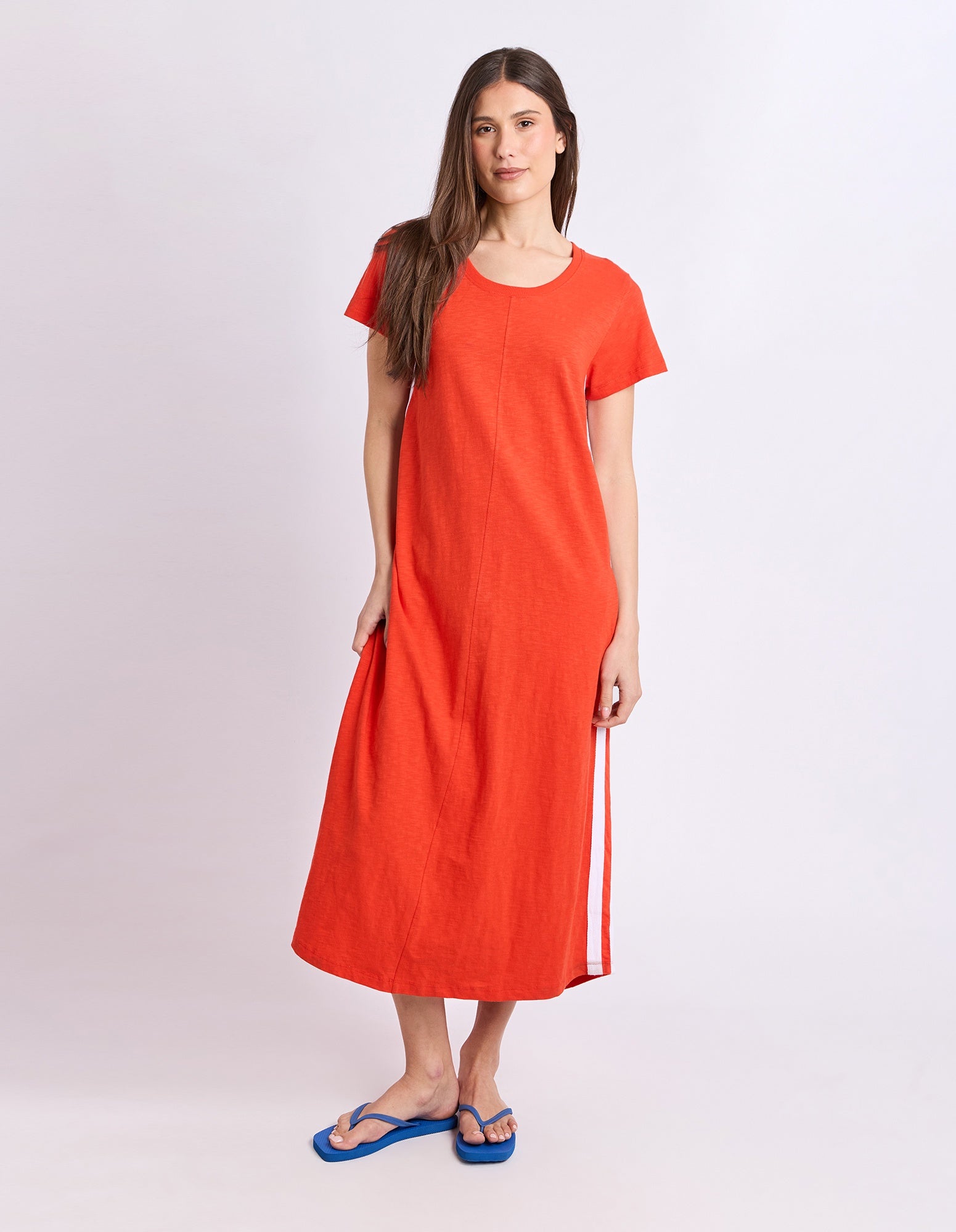 A woman modeling a bright orange short-sleeve midi dress with a white side stripe, paired with blue flip-flop sandals, against a light gray studio background.
