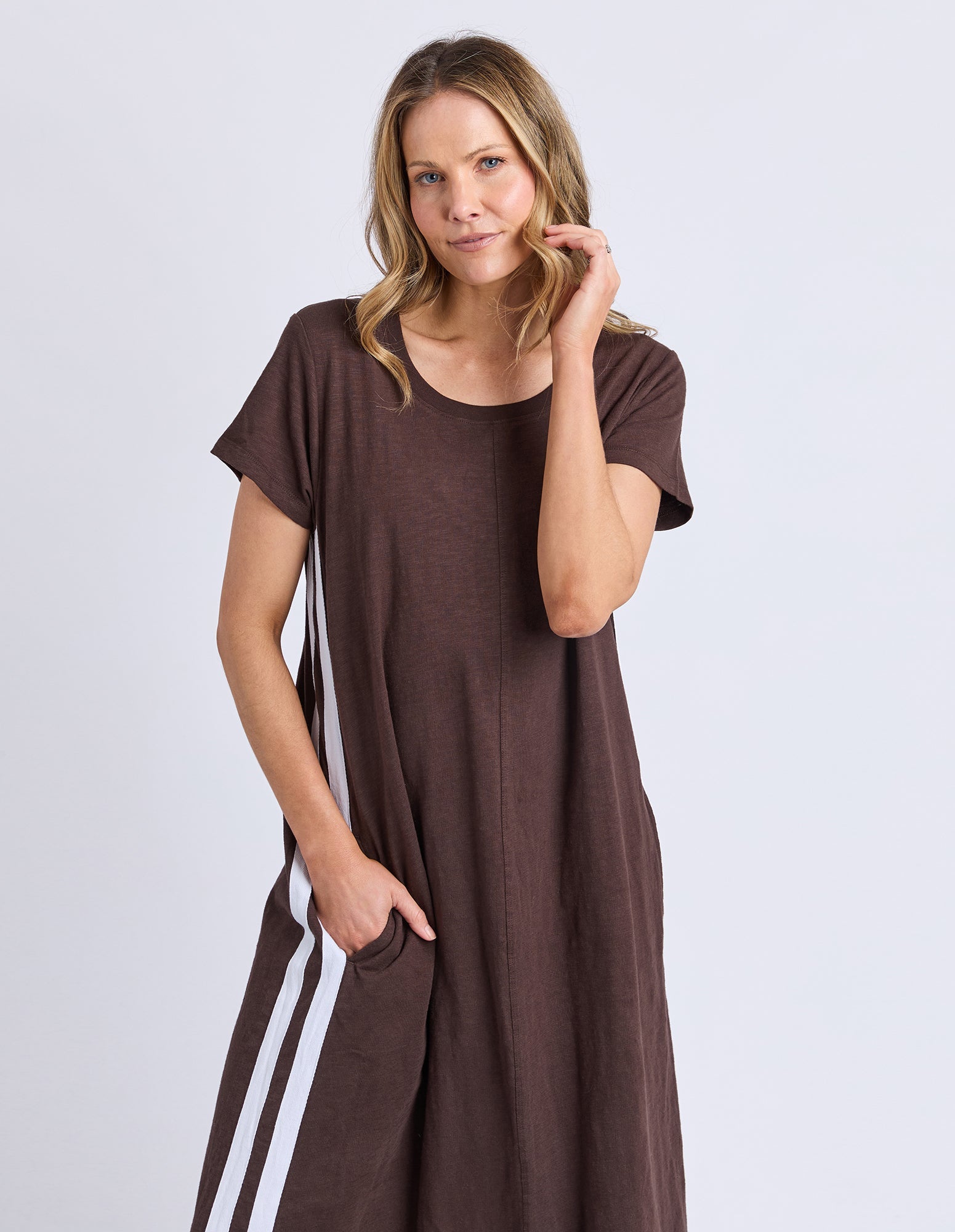 A woman modeling a brown short-sleeve maxi dress with white double stripes down the sides, set against a plain light gray studio background.