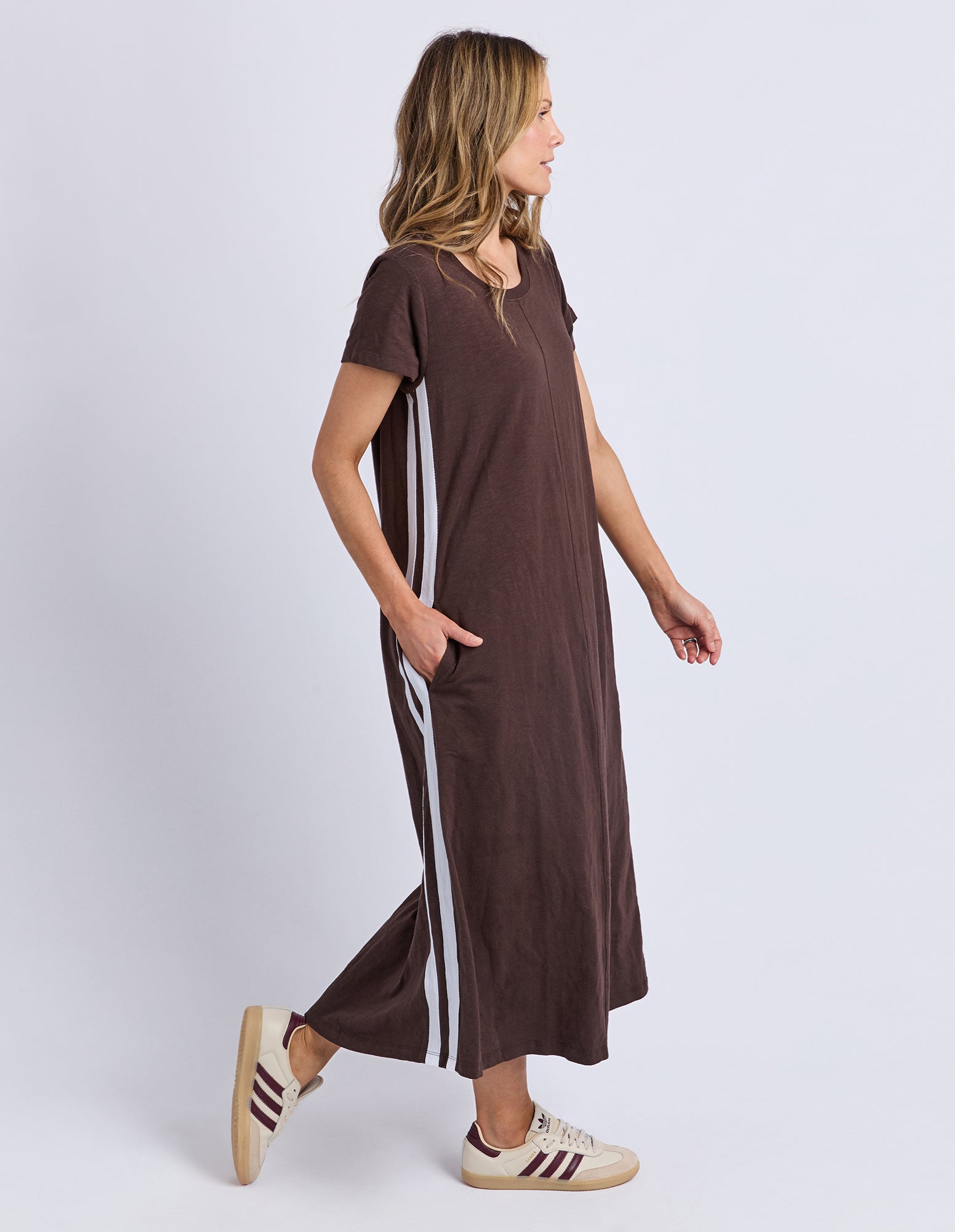 A woman wearing a brown short-sleeve maxi dress with white side stripes, paired with beige sneakers, shown from a side profile against a light gray studio background.