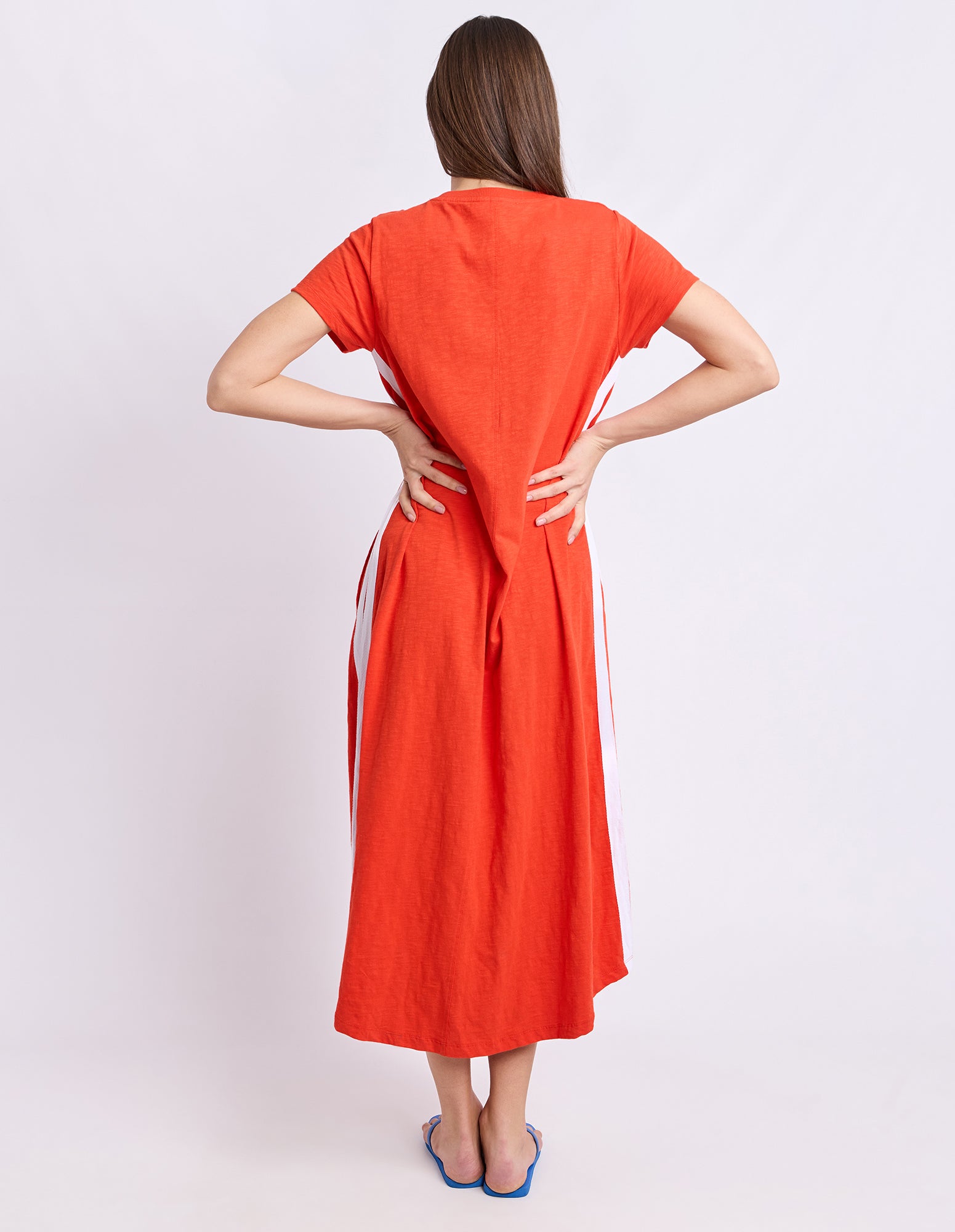 A woman viewed from the back wearing a bright orange short-sleeve midi dress with white side panels and blue sandals, standing against a light gray studio background.