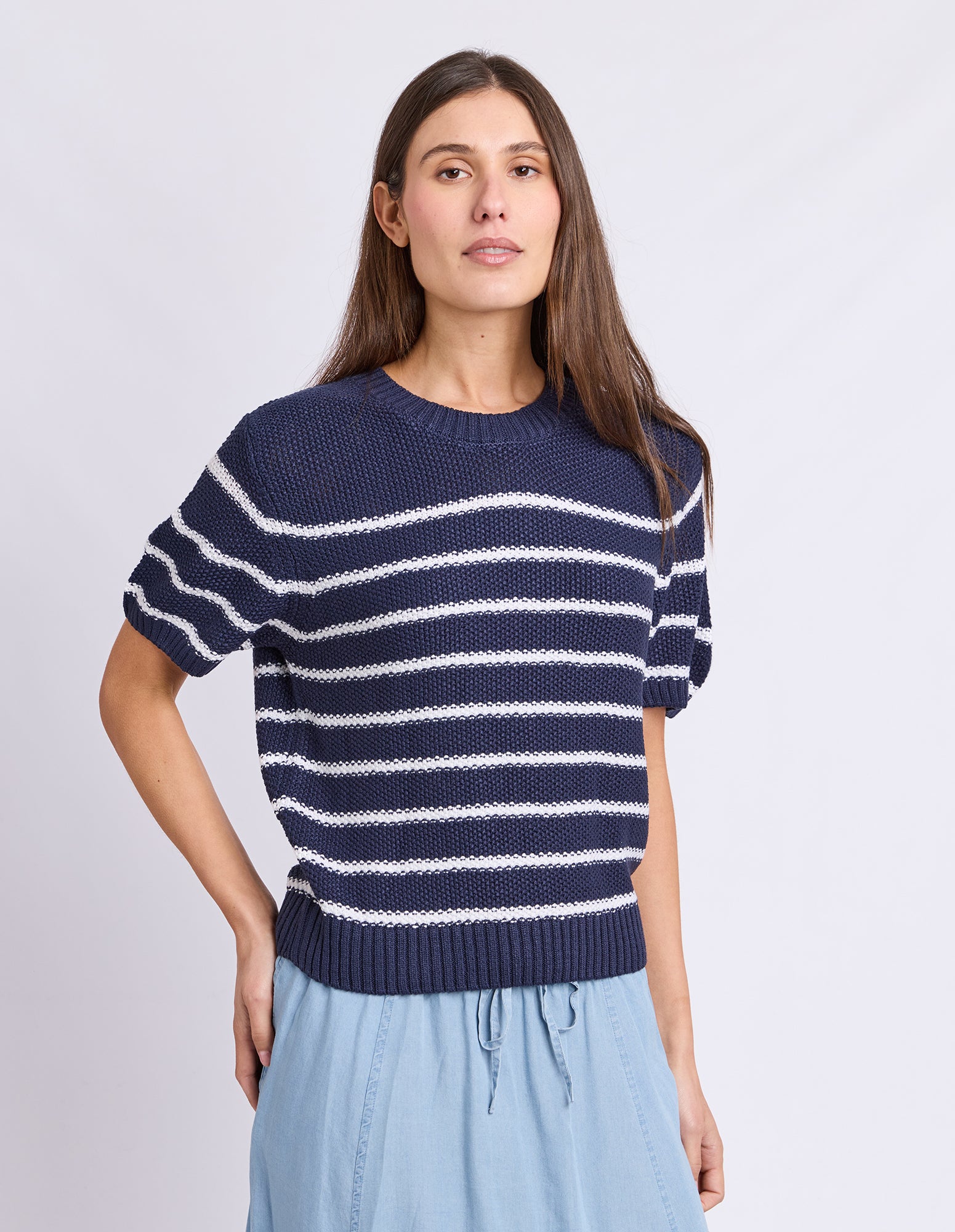 A woman wearing a navy blue short-sleeve knit sweater with white horizontal stripes and light blue skirt, standing against a light gray studio background.