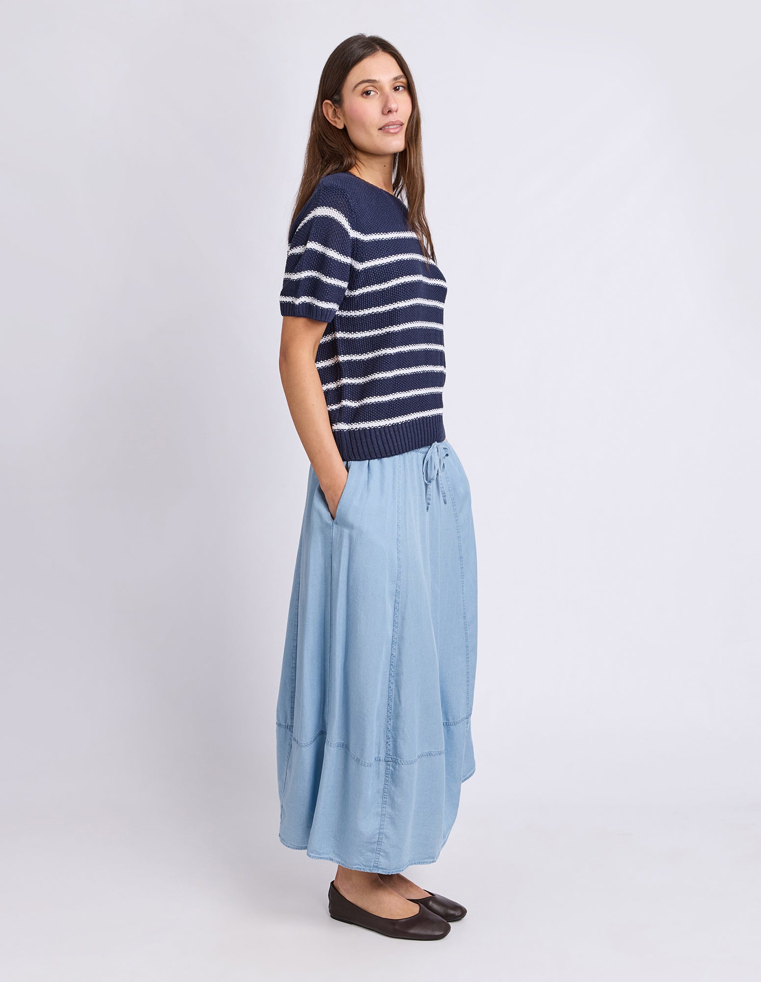 A woman modeling a short-sleeve knit top with navy and white horizontal stripes paired with a light blue maxi skirt and dark brown flats, set against a plain light gray studio background.