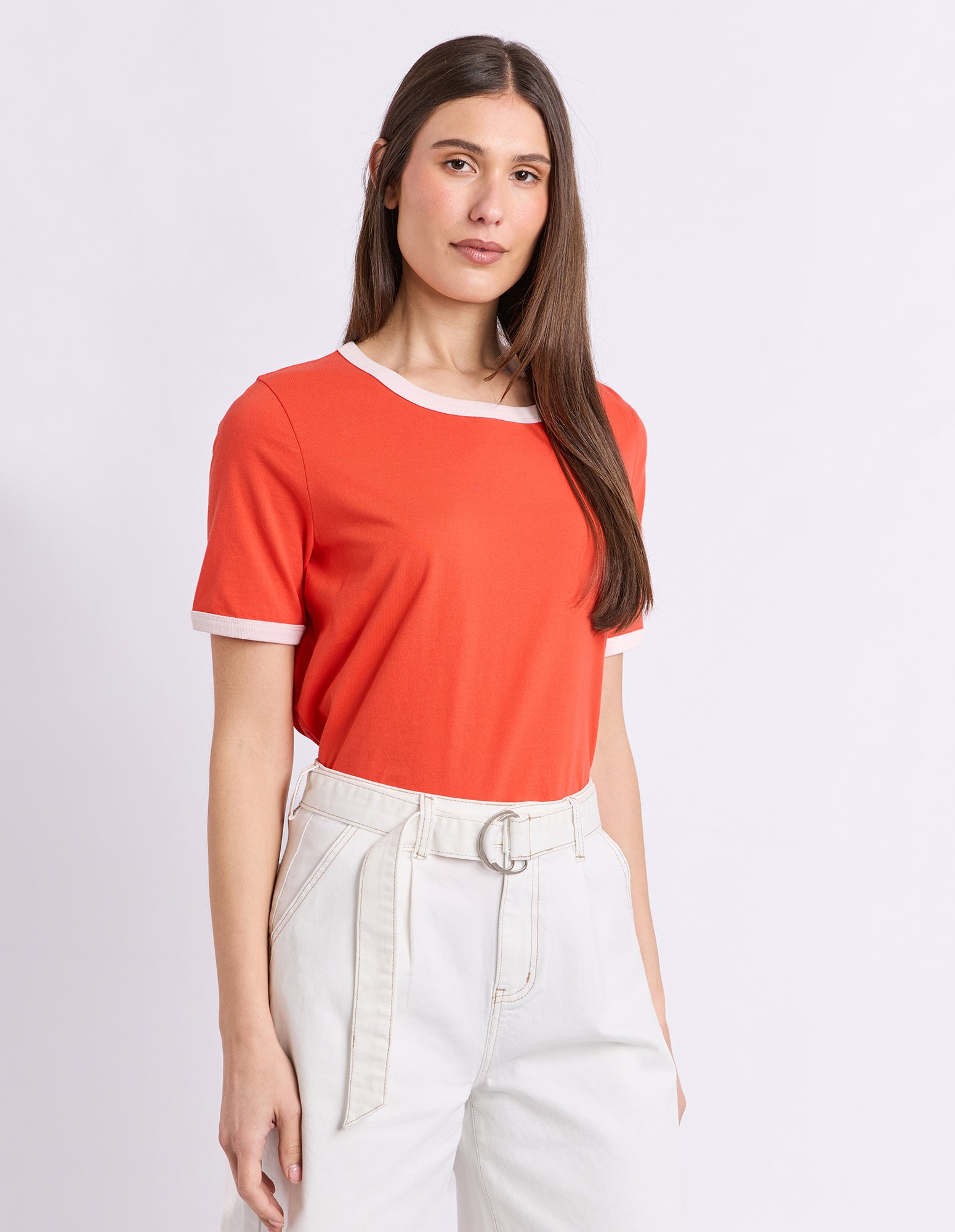 A woman modeling a short-sleeve ringer t-shirt in a vibrant orange-red color with contrasting white trim at the crew neck and sleeve cuffs, paired with high-waisted off-white trousers.