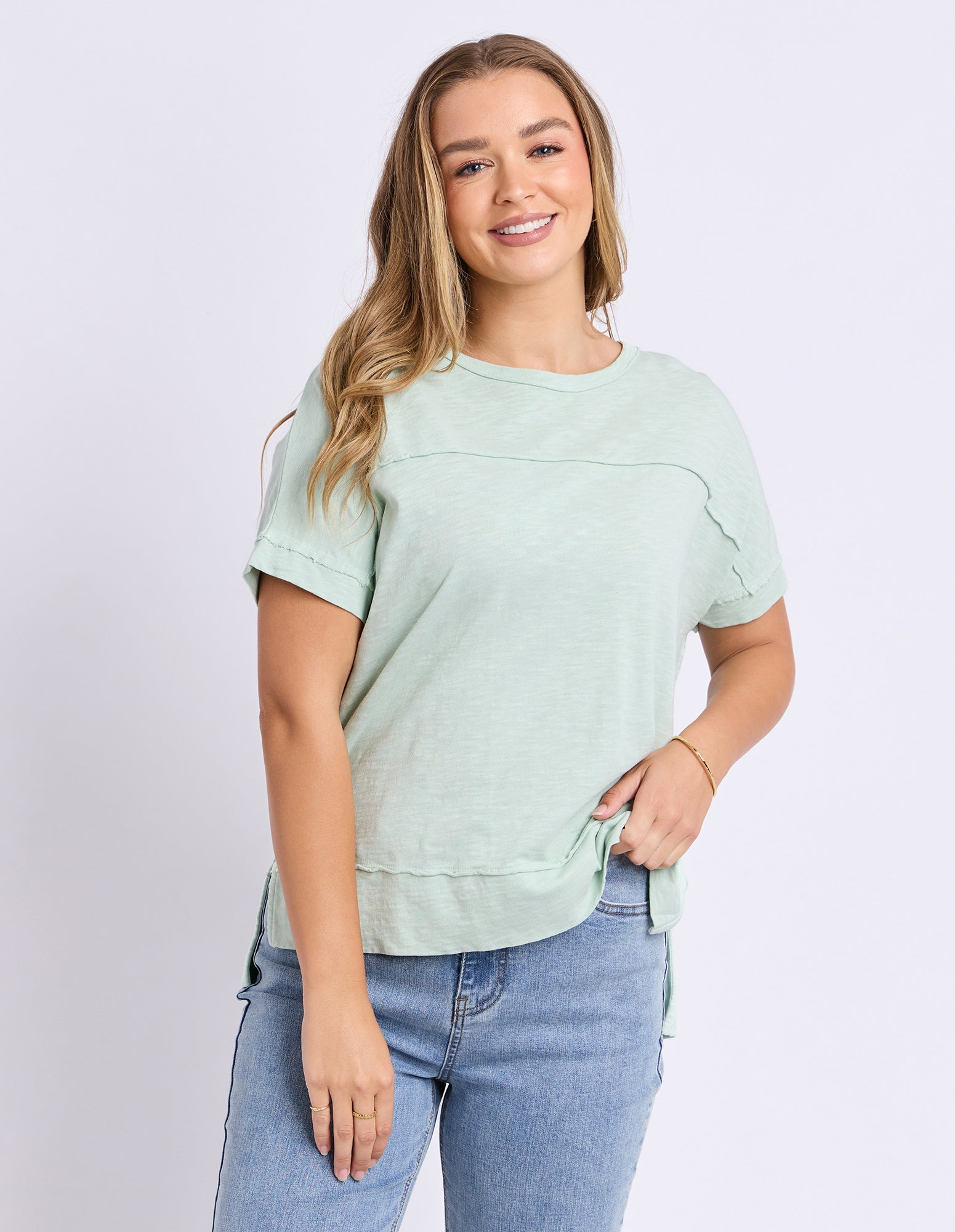 A woman modeling a light sage green short-sleeve top with shoulder cutouts and ruffle details, paired with blue jeans, against a light gray studio background.
