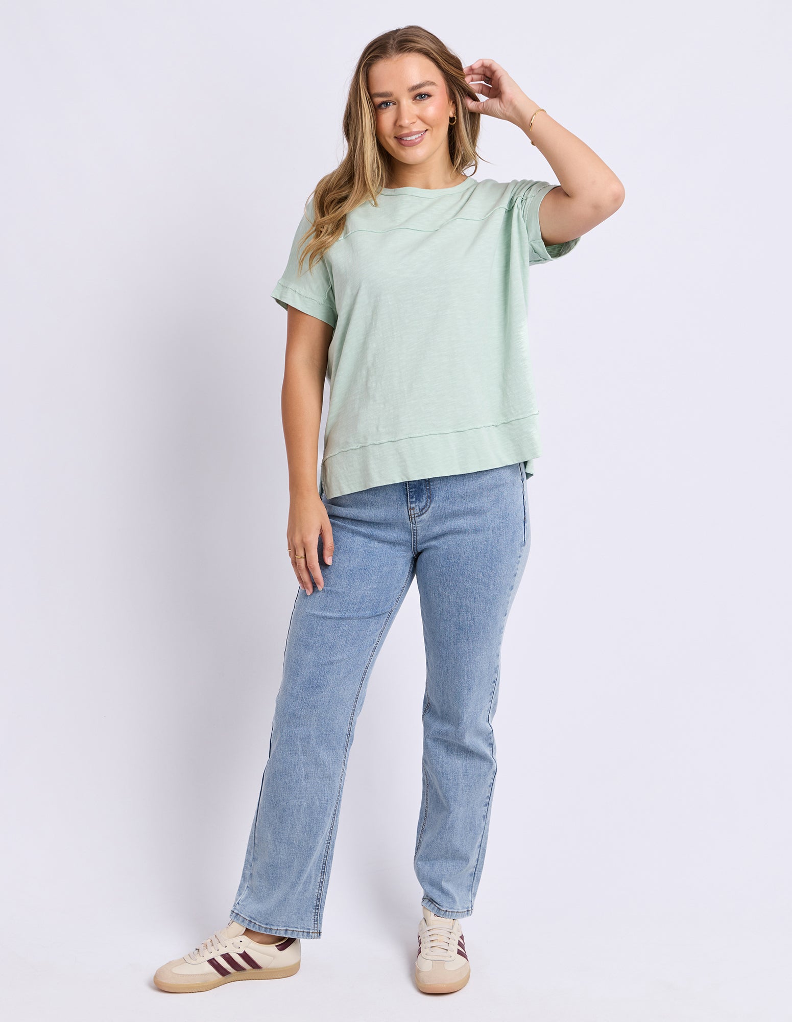 A woman modeling a light sage green short-sleeve crewneck sweatshirt and light-wash blue jeans, paired with beige sneakers, against a plain light gray studio background.