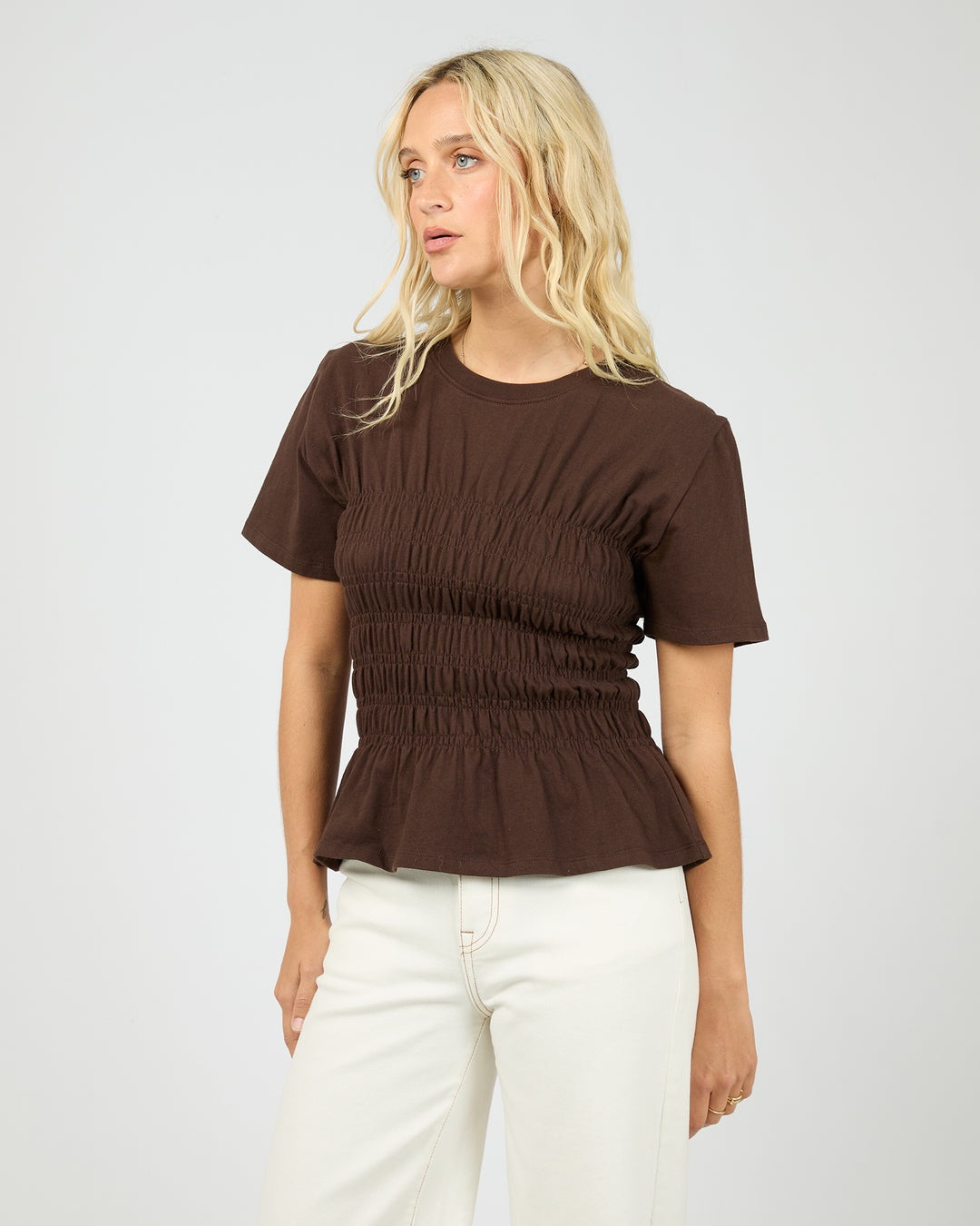 Woman wearing a brown ruched top and white pants on a plain background