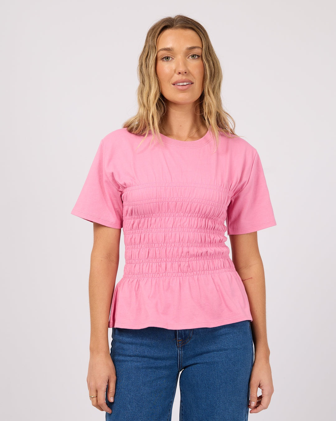 A woman wearing a pink short-sleeve smocked blouse and blue jeans, standing against a light gray studio background.