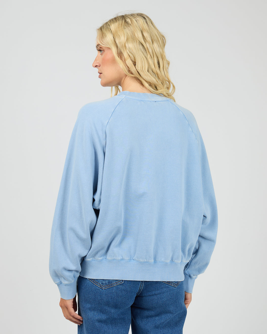 A woman with blonde hair is shown from the back wearing a light blue crewneck sweatshirt and blue jeans against a plain light gray background.