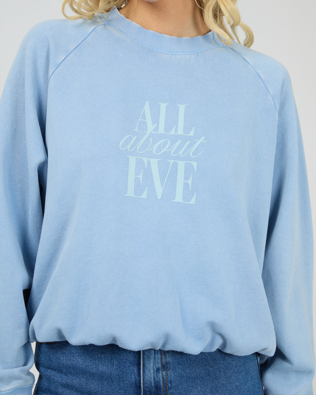 A person wearing a light blue crewneck sweatshirt with the text 'ALL about EVE' printed in tonal light blue on the front, paired with blue jeans against a plain light background.