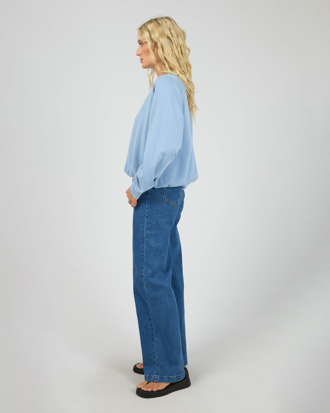 A woman stands in profile wearing a light blue long-sleeve blouse and wide-leg blue jeans against a plain light gray studio background.