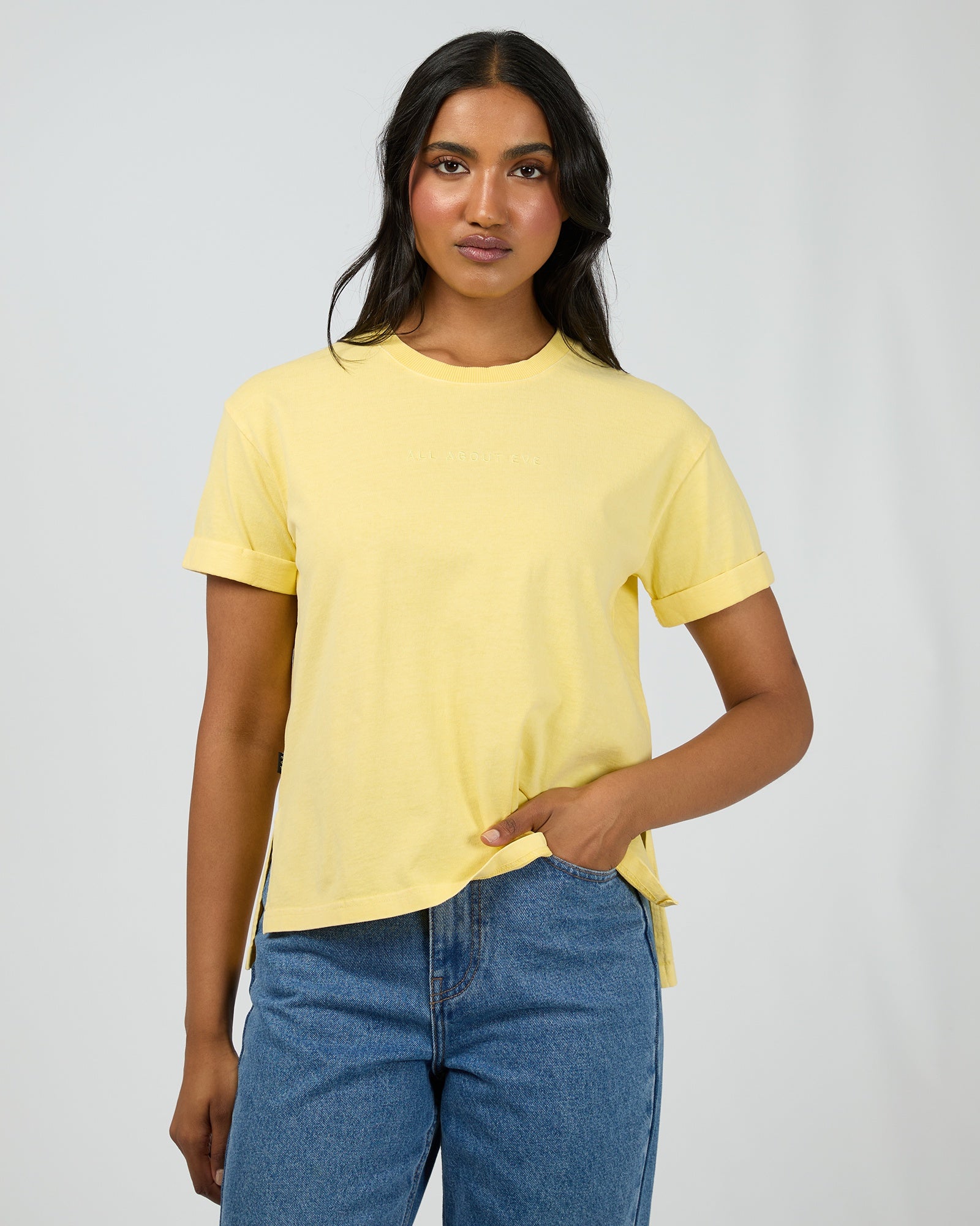 A woman modeling a pale yellow short-sleeve crewneck t-shirt paired with blue jeans, standing against a light gray studio background.