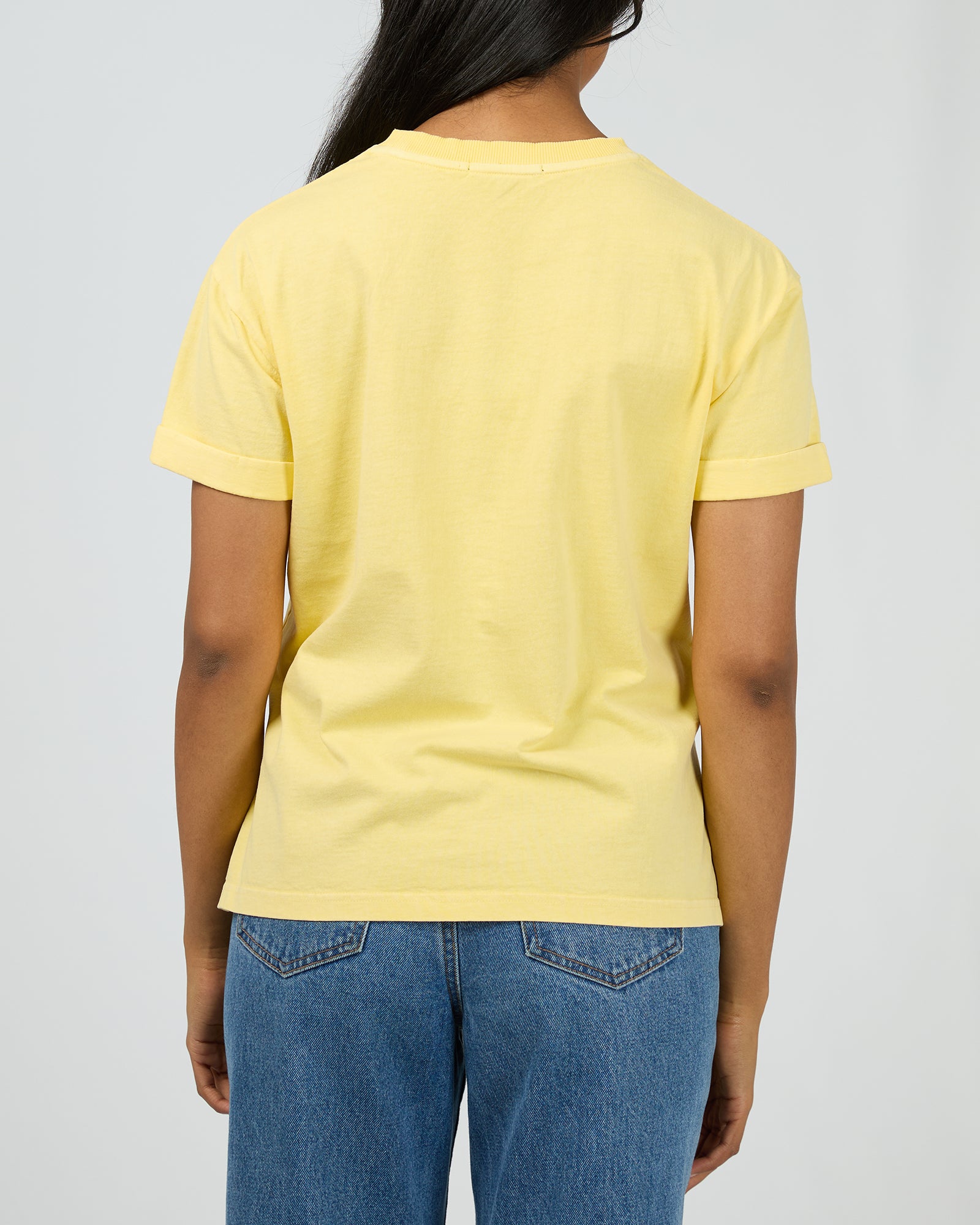 A person shown from the back wearing a pale yellow short-sleeve crewneck T-shirt and blue jeans against a light gray studio background.