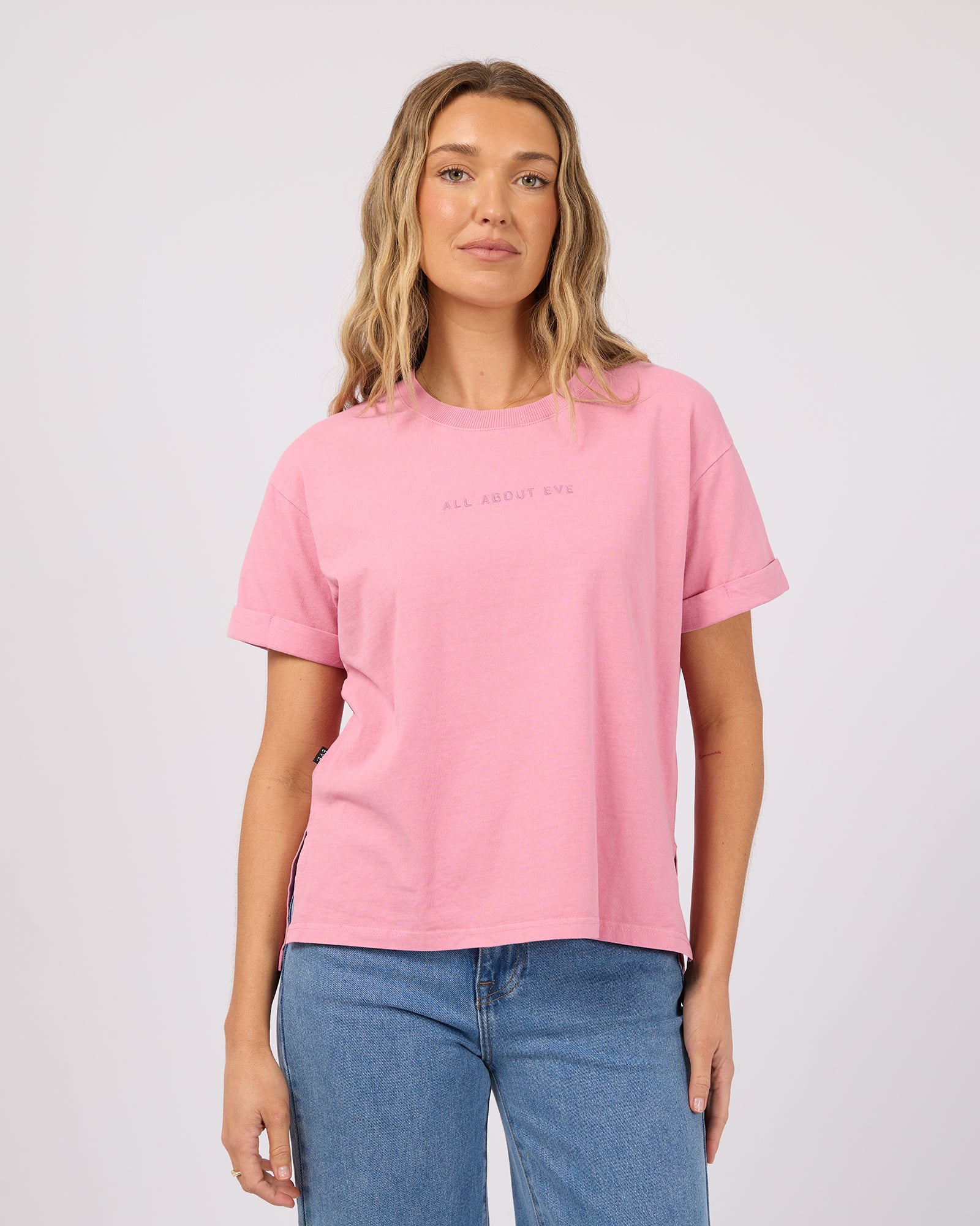 A woman wearing a light pink short-sleeve t-shirt with 'ALL ABOUT ME.' printed in small dark letters on the chest, paired with blue jeans, against a light gray studio background.