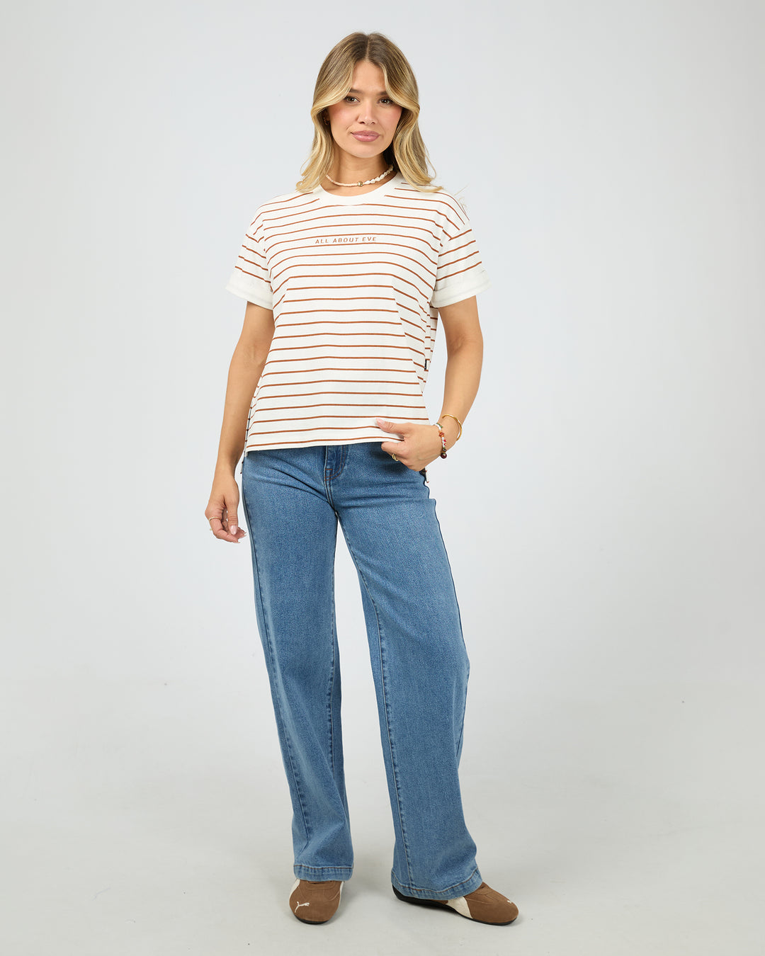 A woman modeling a short-sleeve crewneck t-shirt with thin brown horizontal stripes on an off-white background, paired with blue jeans and brown shoes, against a light gray studio background.